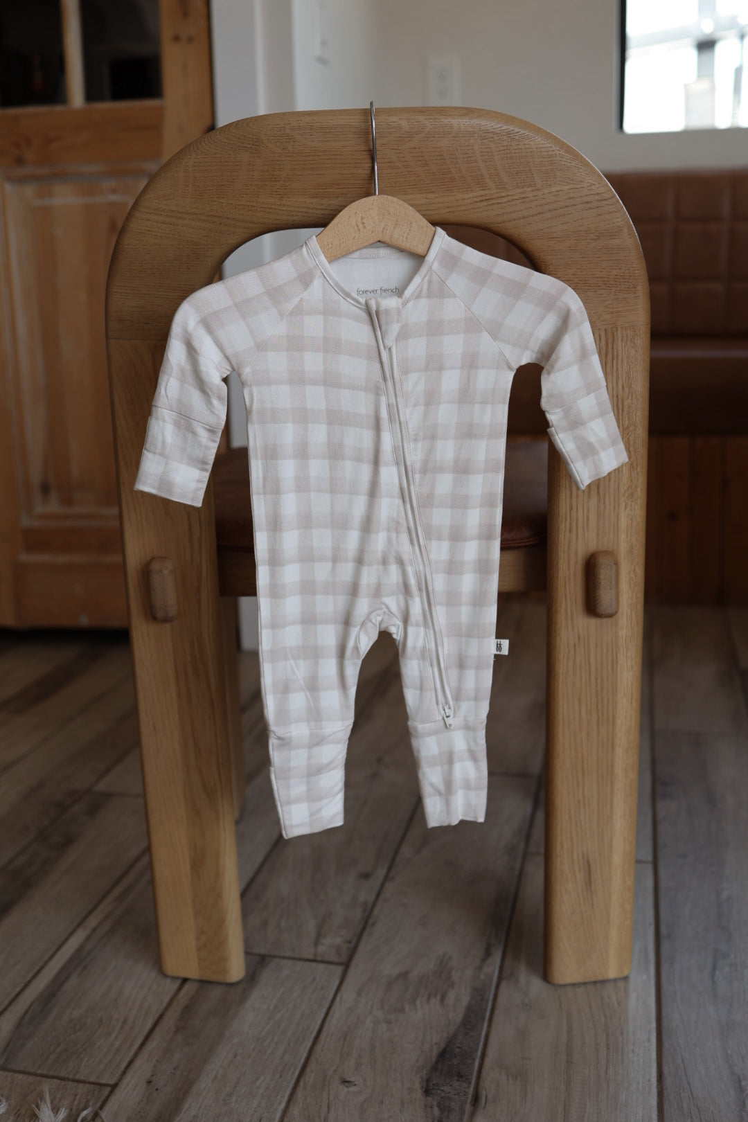 The forever french baby Bamboo Zip Pajamas | Gingham, a beige and white plaid onesie with long sleeves and a zipper front, hangs on a wooden hanger over a chair in a cozy room.