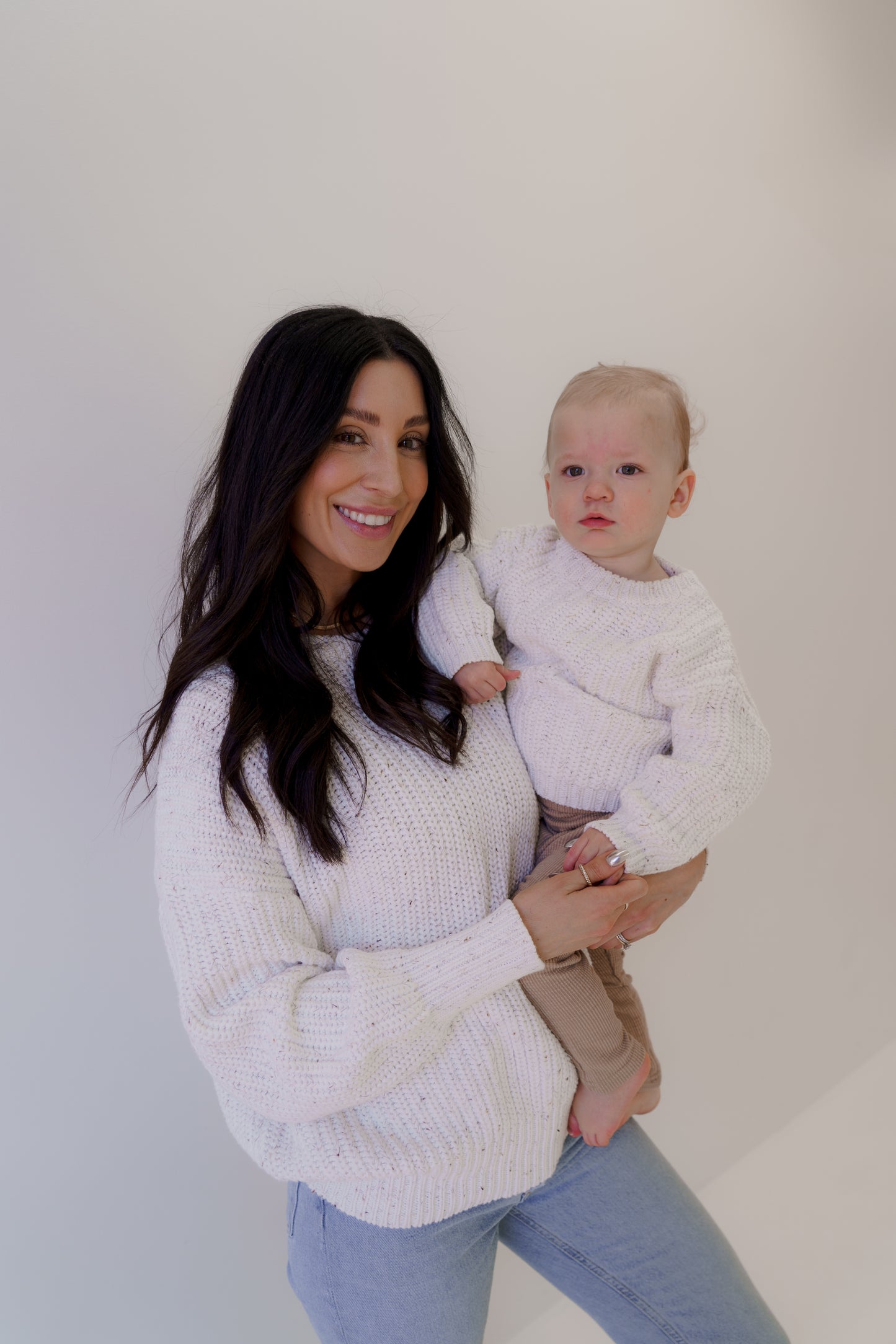A woman with long dark hair smiles while holding her baby, both dressed in white "Child Knit Sweaters" from forever french baby. The baby's expression is neutral, and they stand against a simple white backdrop.