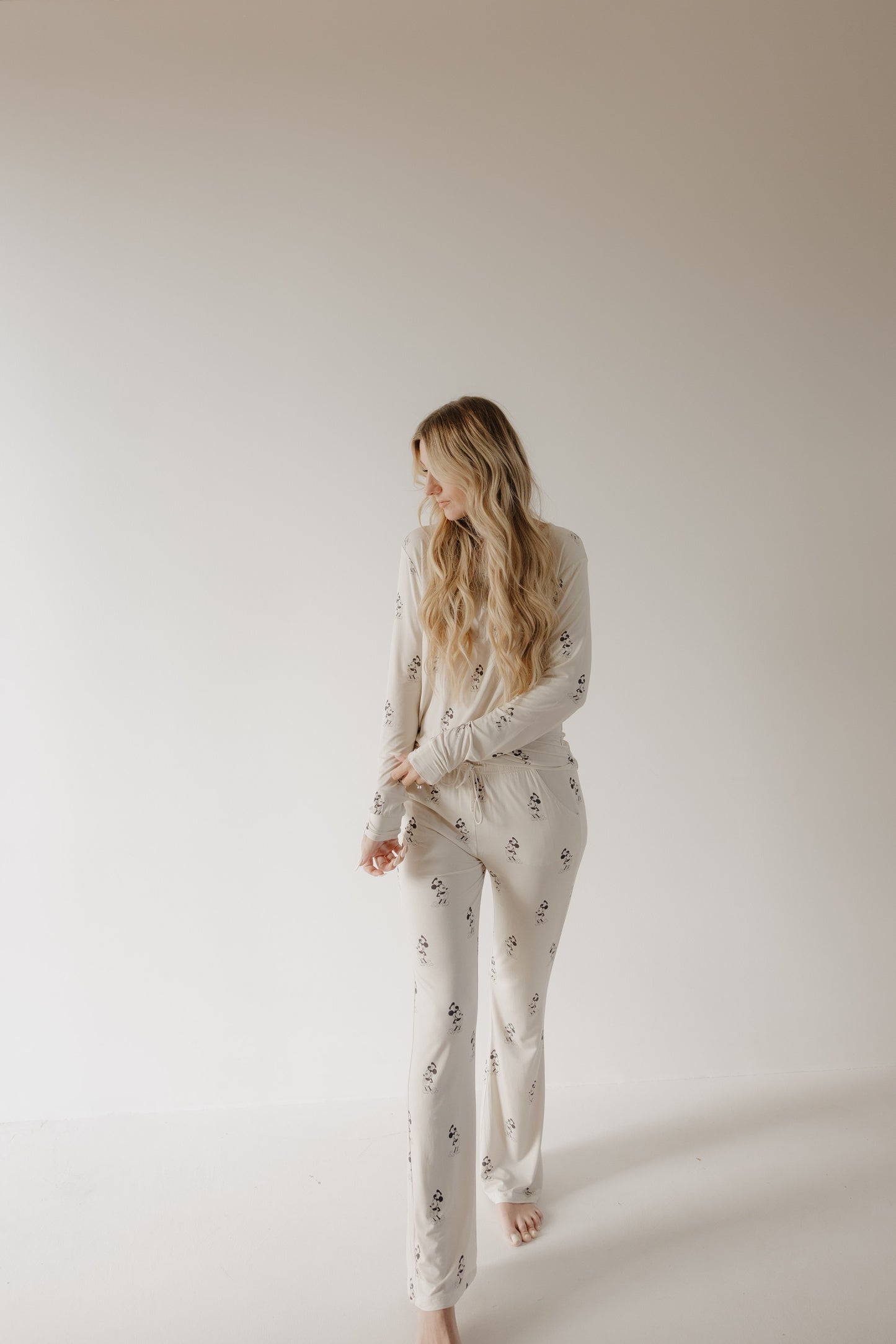 A woman with long, wavy blonde hair stands barefoot against a white background, wearing forever french baby Women’s Bamboo Pajama | All Aboard!, which features a light color and subtle dark pattern. She looks left, arms relaxed.