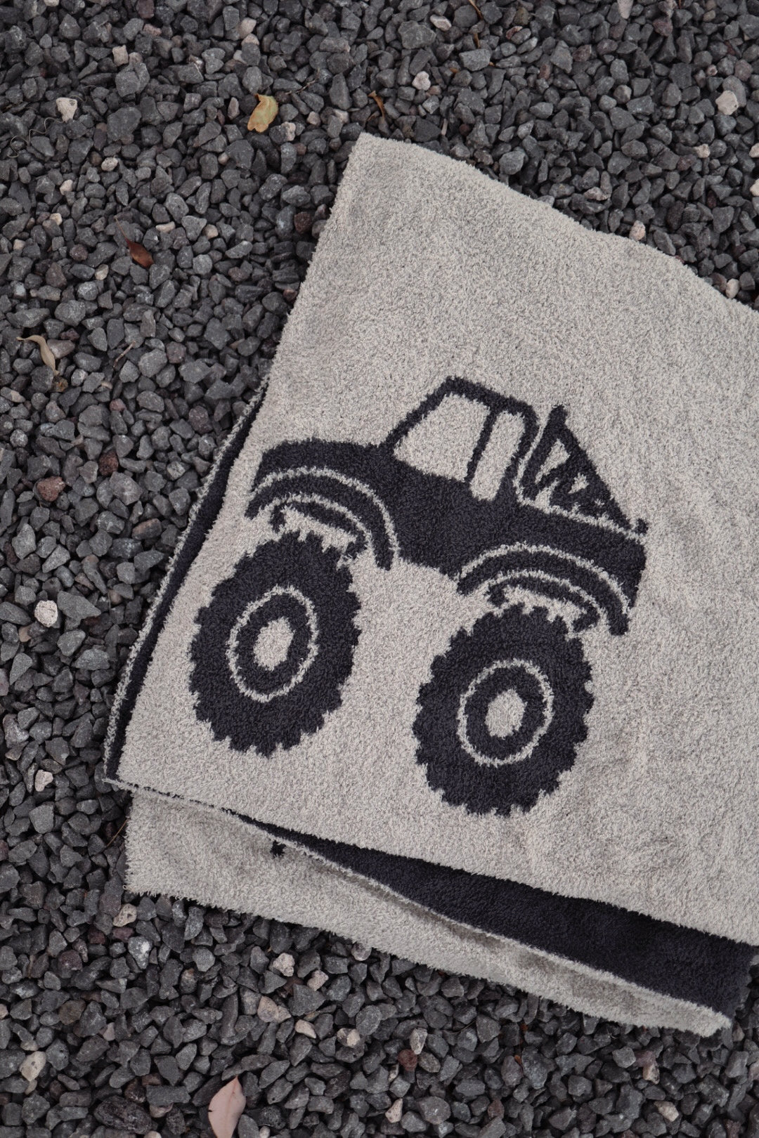 The forever french baby Plush Blanket | Monster Truck, featuring a black outlined monster truck design on light gray microfiber, is displayed on top of a towel over small dark gravel stones.