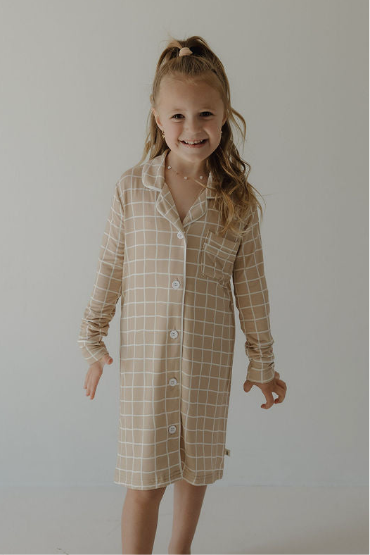 A smiling young girl with long, wavy blonde hair wears the forever french baby Toddler Bamboo Sleeping Dress | Grid, featuring a soft beige and white checkered design, standing against a plain light background.