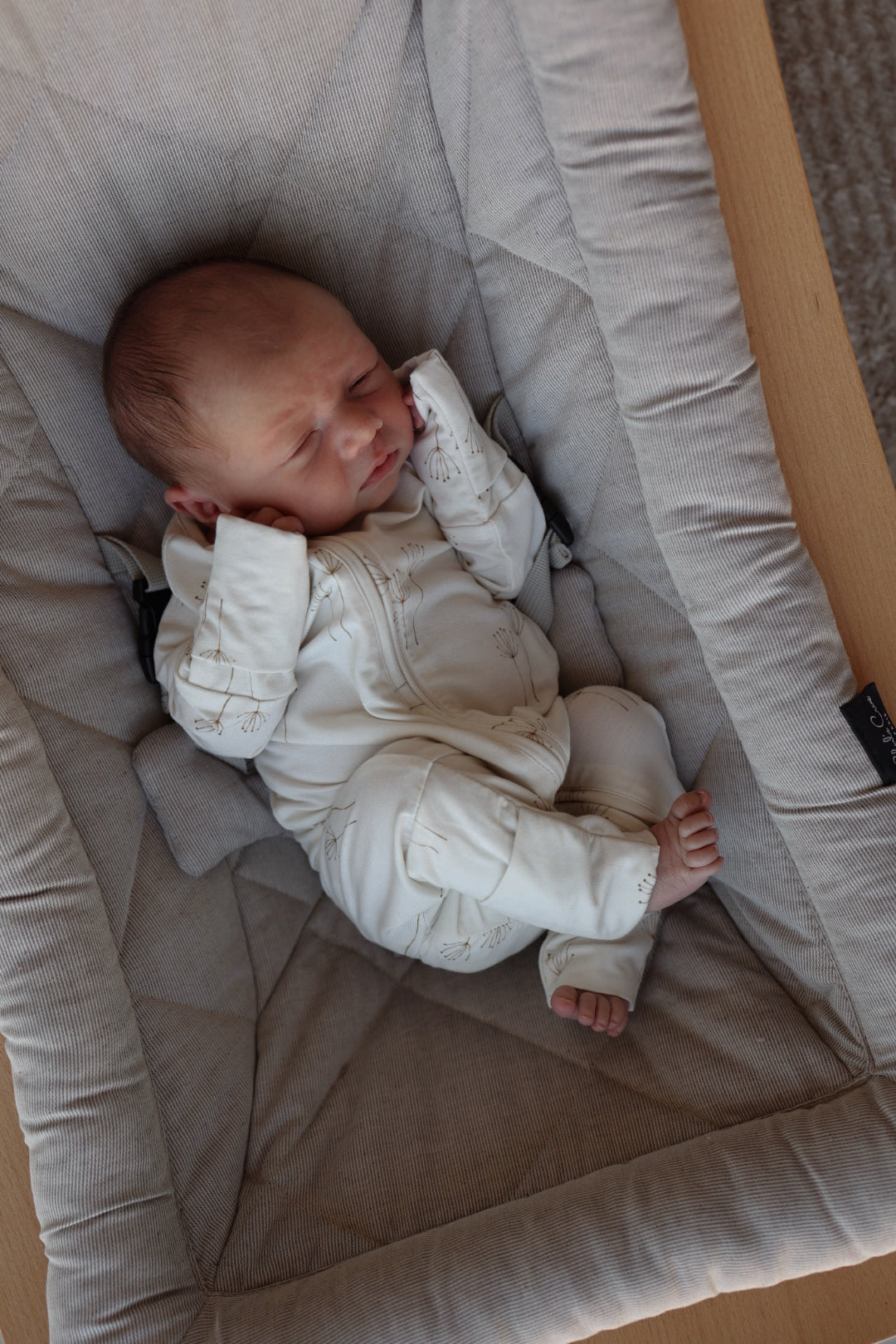 A newborn sleeps peacefully in a cushioned light-gray seat, dressed in forever french baby Bamboo Zip Pajamas | Dandelion, with hands near their face and feet relaxed. The cozy seat rests on a wooden surface.