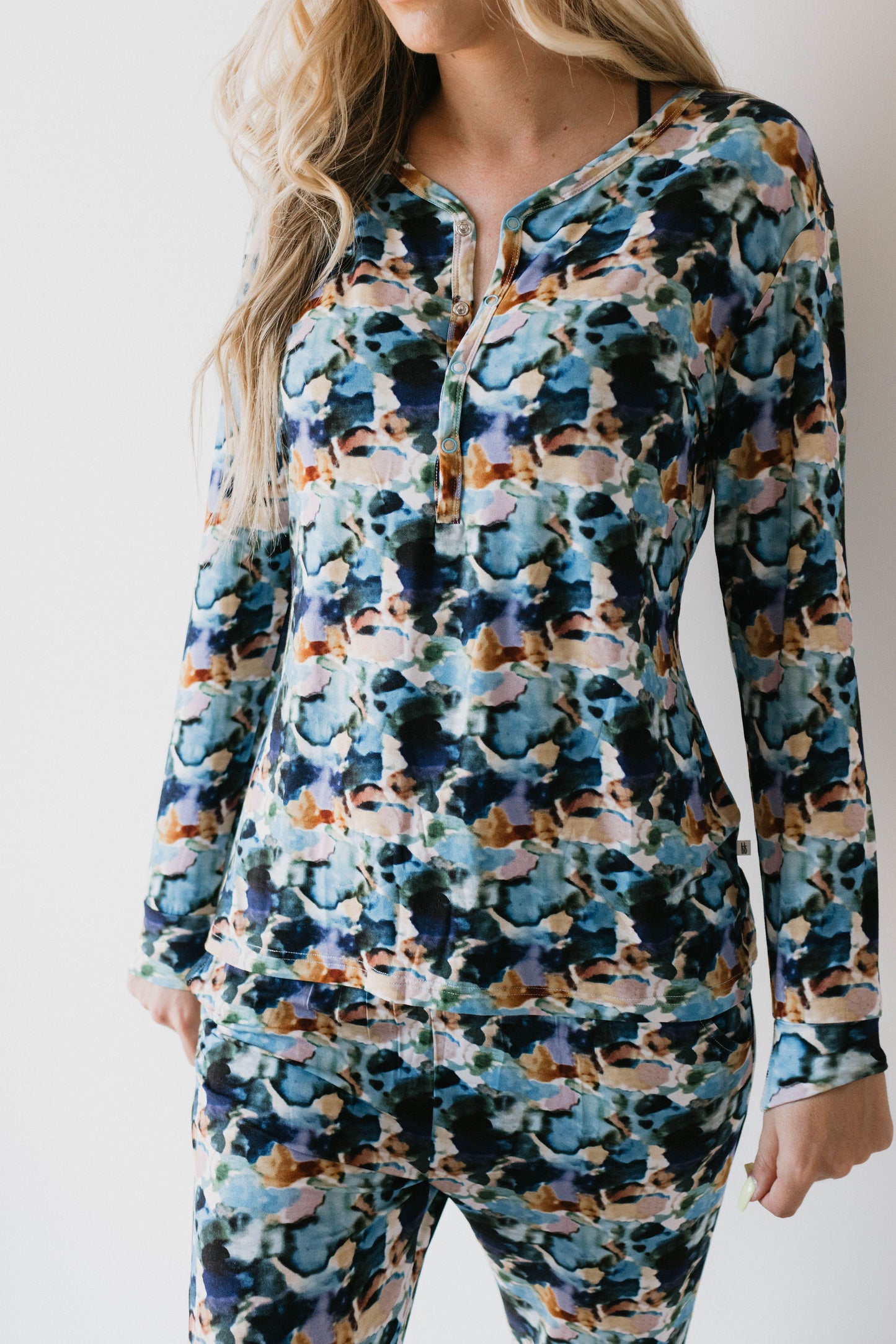 A person is wearing the Women's Bamboo Pajamas in the Charli Print from forever french baby, which feature a colorful, abstract pattern with shades of blue, black, green, and tan. The long-sleeve top has a zipper at the neckline and matches the pants. The person has long, wavy blonde hair, with only their torso and upper legs visible.
