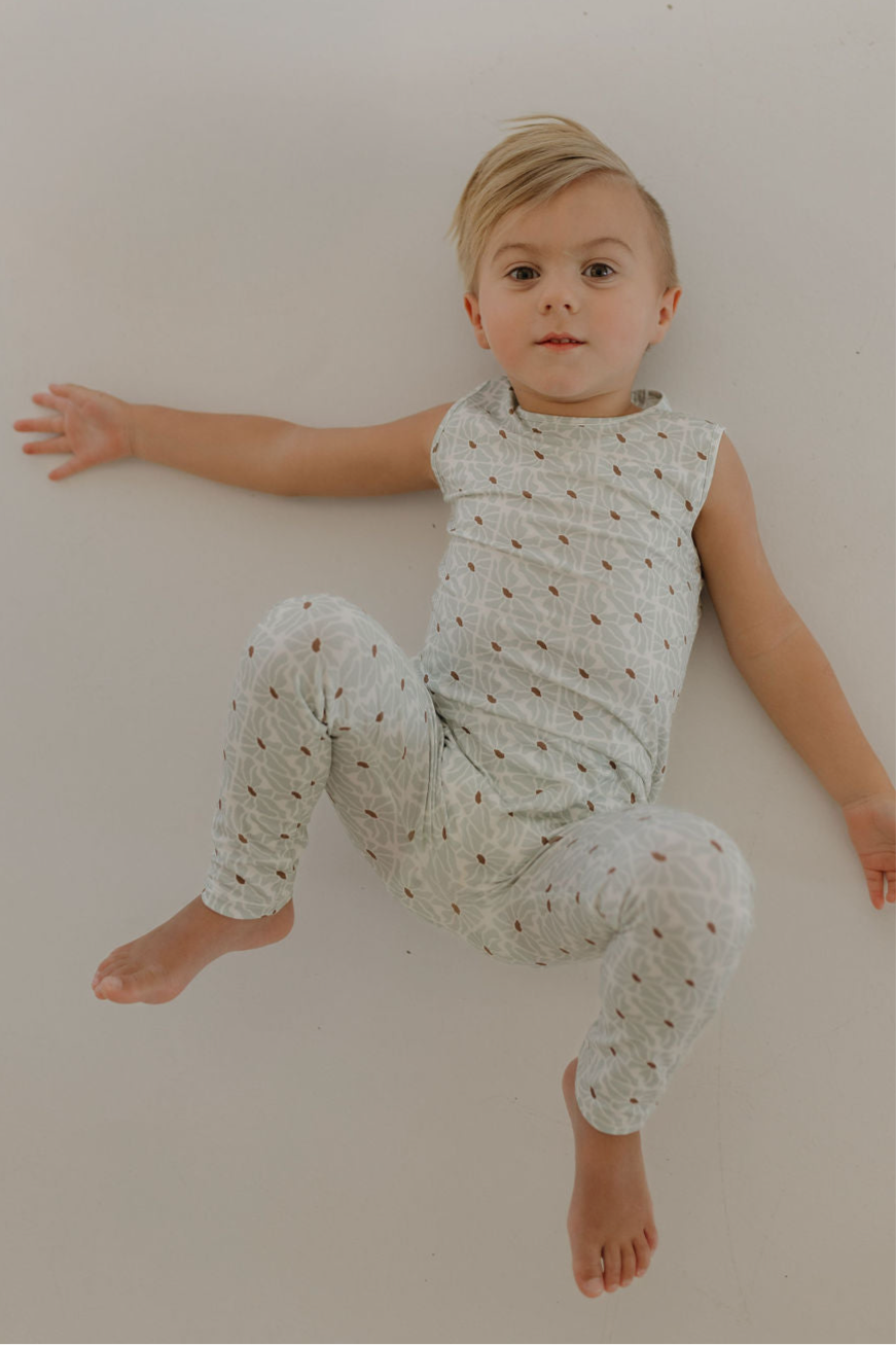 A young child in the forever french baby Harem Bamboo Romper | Coast, made of breathable bamboo, lies on a white surface in light geometric-patterned pajamas, stretching out an arm and looking upward in comfort.