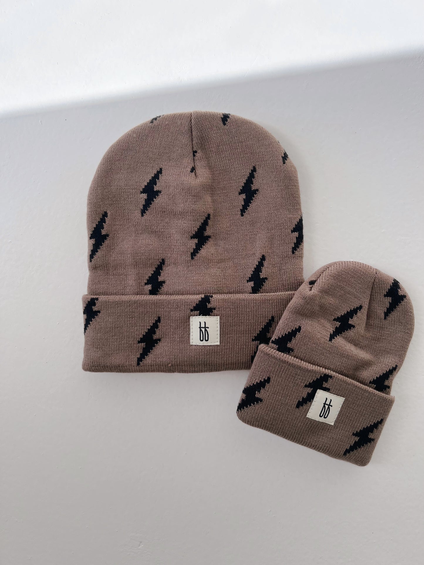 Two ff Knit Beanies from Forever French Baby, featuring a classic ribbed design and black lightning bolt patterns, are placed on a white surface. One beanie is larger while the other is smaller. Both have a small white tag displaying the brand's logo with two lowercase "b" letters.