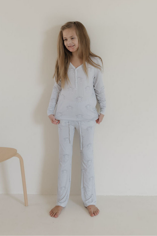 A smiling young girl with long blonde hair stands barefoot against a light wall, wearing forever french baby’s Youth Flare Bamboo Pajamas in the Howdy print. A wooden chair sits partially visible on the left as she looks off to the side.