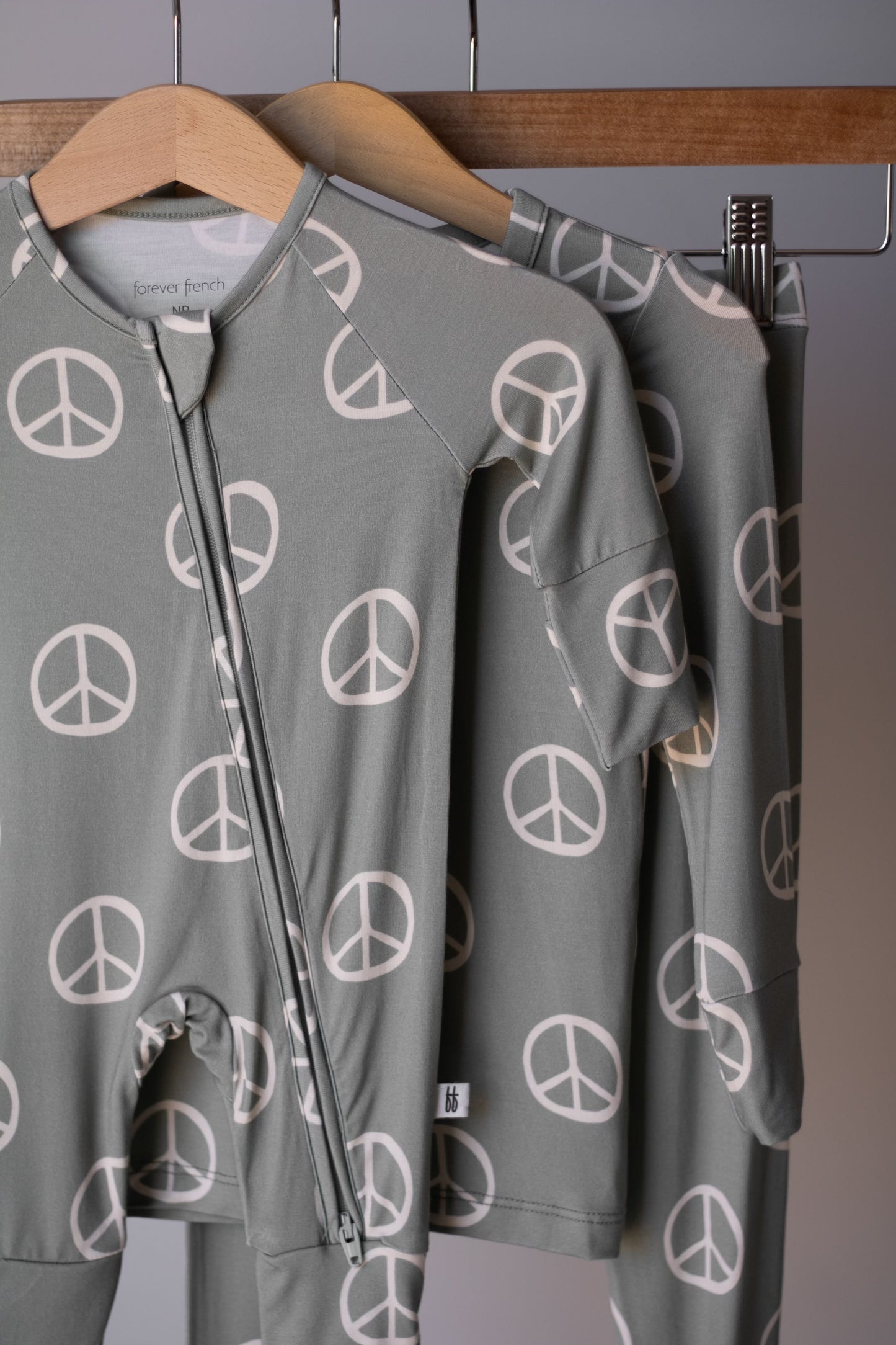 Two sage green "Bamboo Two Piece Pajamas | Peace" by forever french baby, featuring white peace signs, hang on wooden hangers. These soft, hypoallergenic children's pajamas include a long-sleeve top and matching pants.