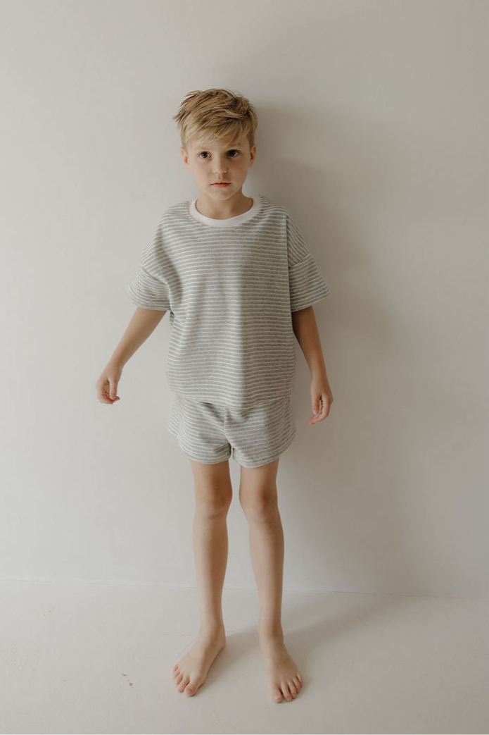 A young blonde boy stands barefoot against a plain white wall, wearing the forever french baby Child Terry Short Set in Grey Stripe. He faces the camera with a neutral expression.