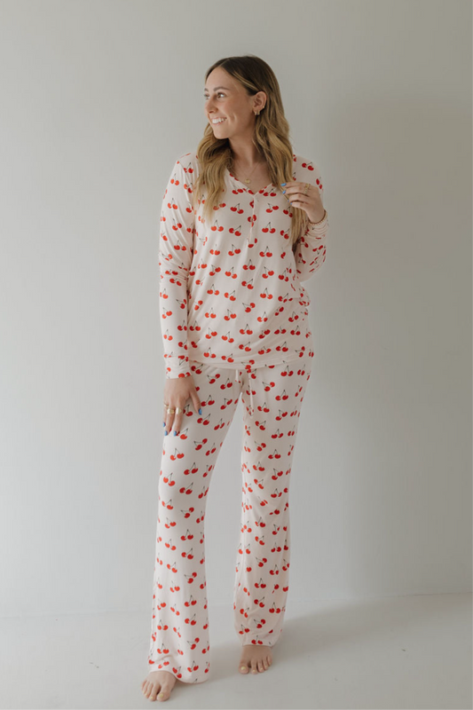 A woman stands barefoot against a light background, smiling to the side. She wears the forever french baby Women's Bamboo Pajama | Cherry Orchard set in cream with a red cherry print, made from breathable bamboo fabric.