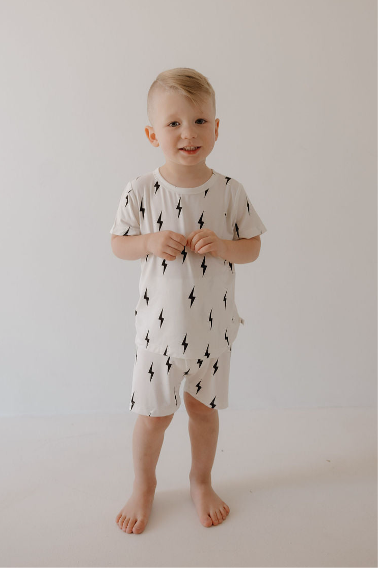 A young child with light hair smiles barefoot against a plain background, wearing the forever french baby Bamboo Short Set in White & Black Lightning Bolt—a breathable sleepwear t-shirt and shorts.