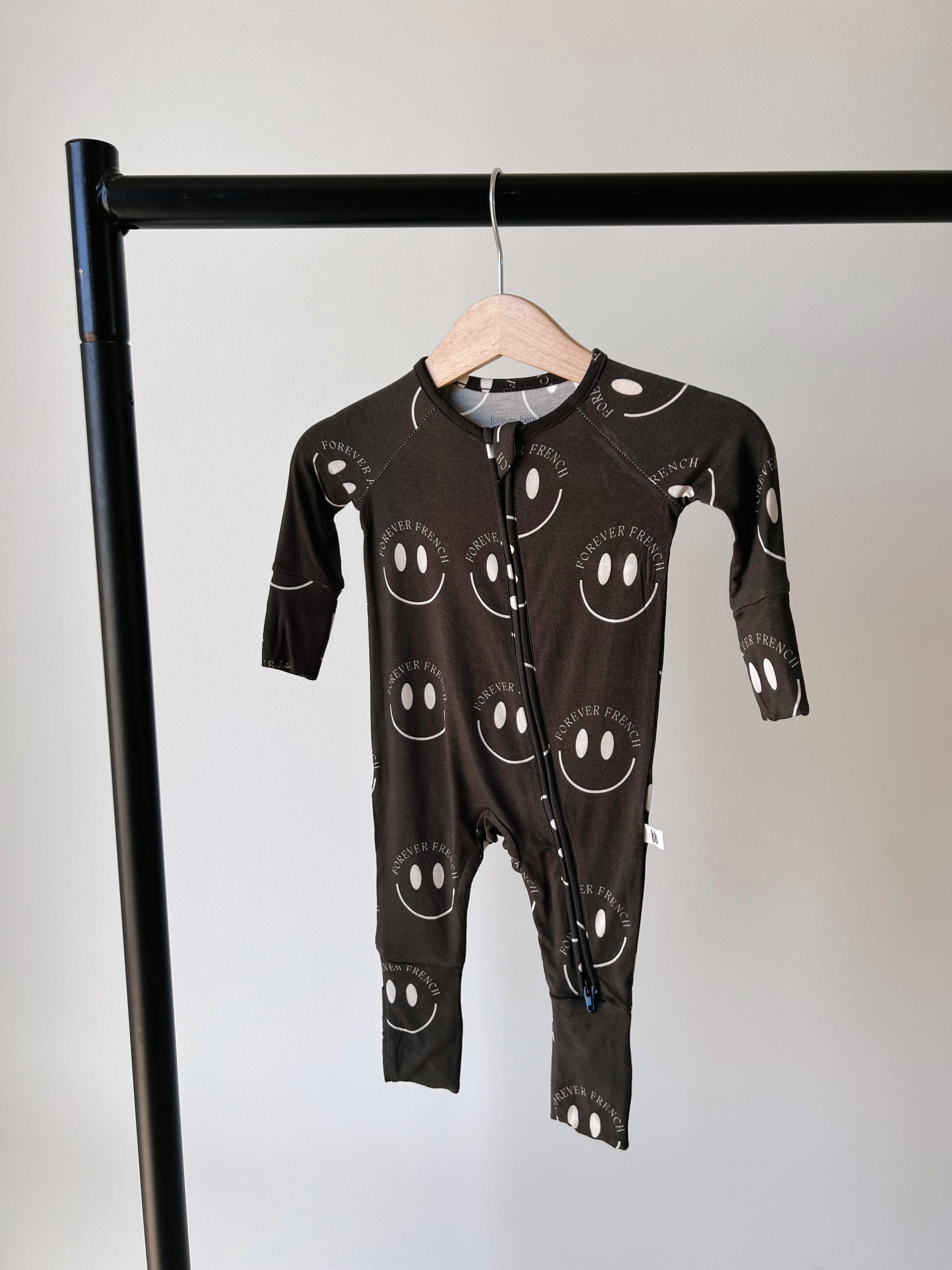 A black "Bamboo Zip Pajamas | Charcoal & White ff Smile" from forever french baby, made from hypo-allergenic fabric with white smiley face patterns and the words "forever french" printed on it. Hanging on a wooden hanger attached to a black metal rod, it features a diagonal zipper from neck to left leg for easy changing.