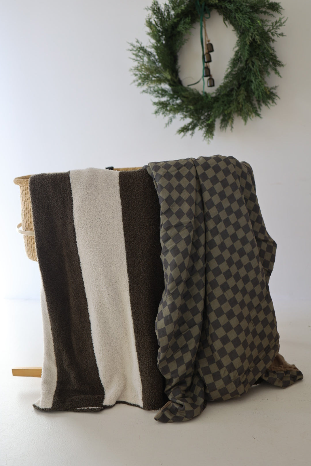 A gray muslin cotton quilt and the Muslin Quilt | Olive Checkerboard by forever french baby drape over a basket; a green wreath with small bells hangs on the wall behind them—perfect cozy nursery bedding.