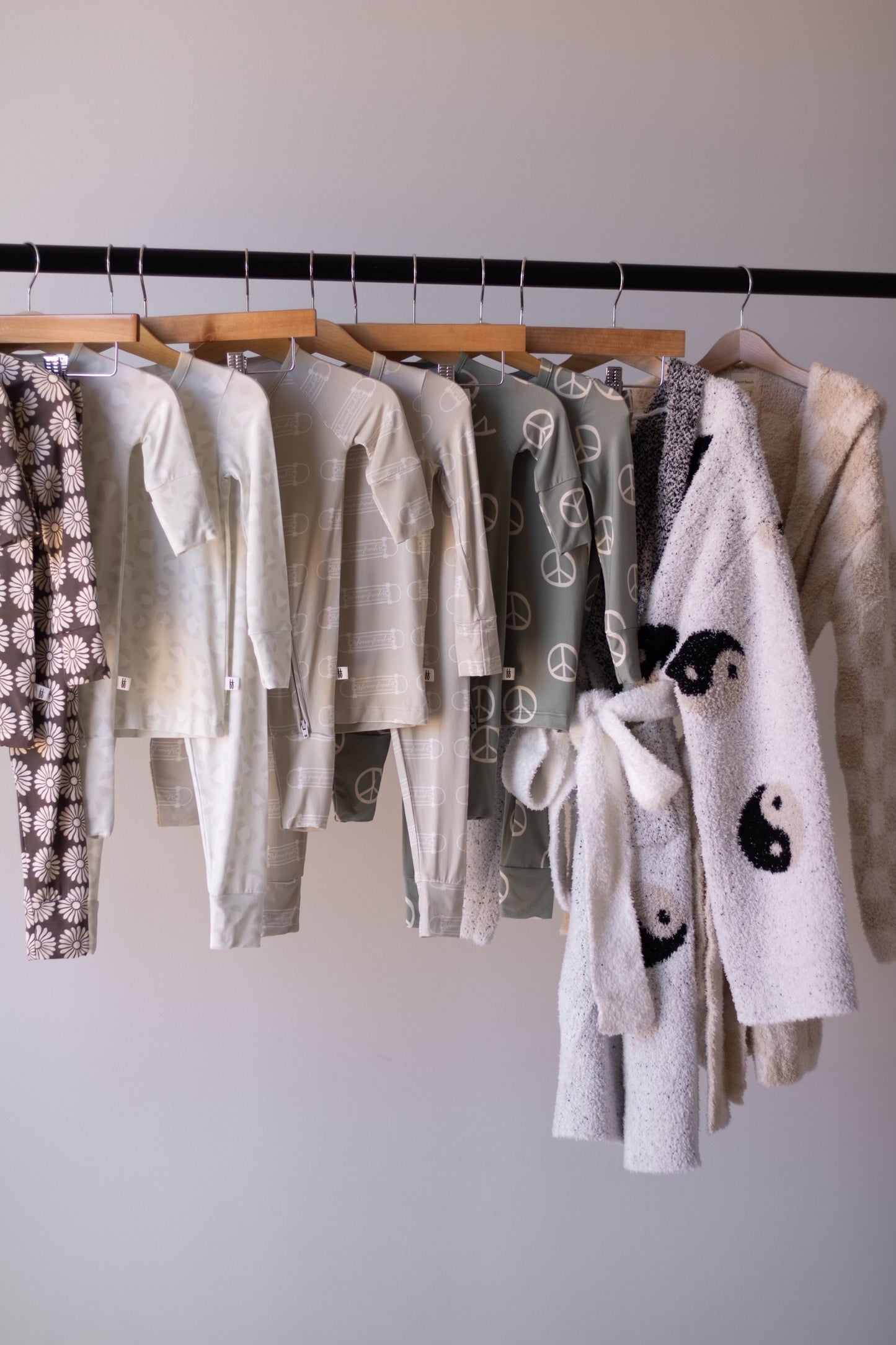 A clothing rack displays forever french baby Bamboo Zip Pajamas | Peace—breathable baby sleepwear in neutral tones with peace signs and other playful patterns—hanging neatly on wooden hangers against a plain wall.