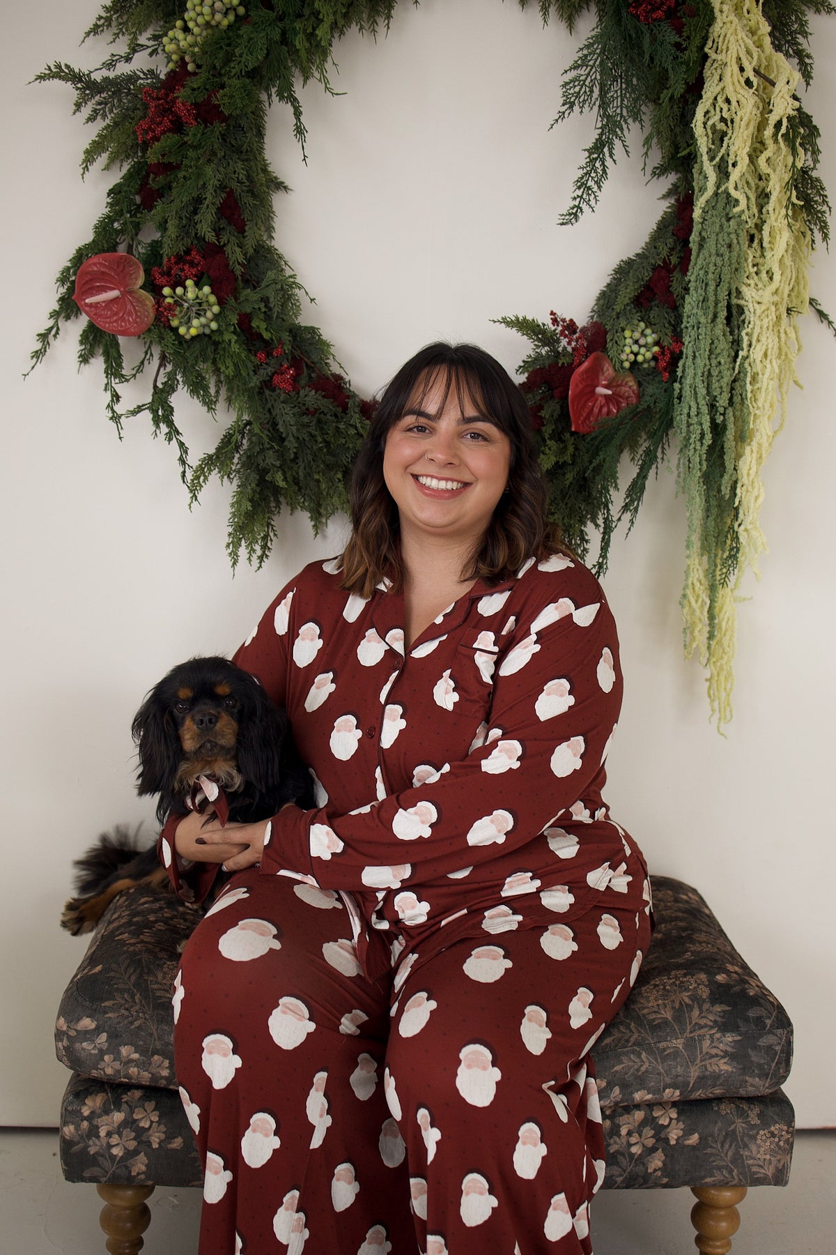 A woman wearing a burgundy Women’s Bamboo Button Up Lounge Set | Vintage Santa sits on an ottoman with a small black and tan dog, posed in front of a holiday wreath.