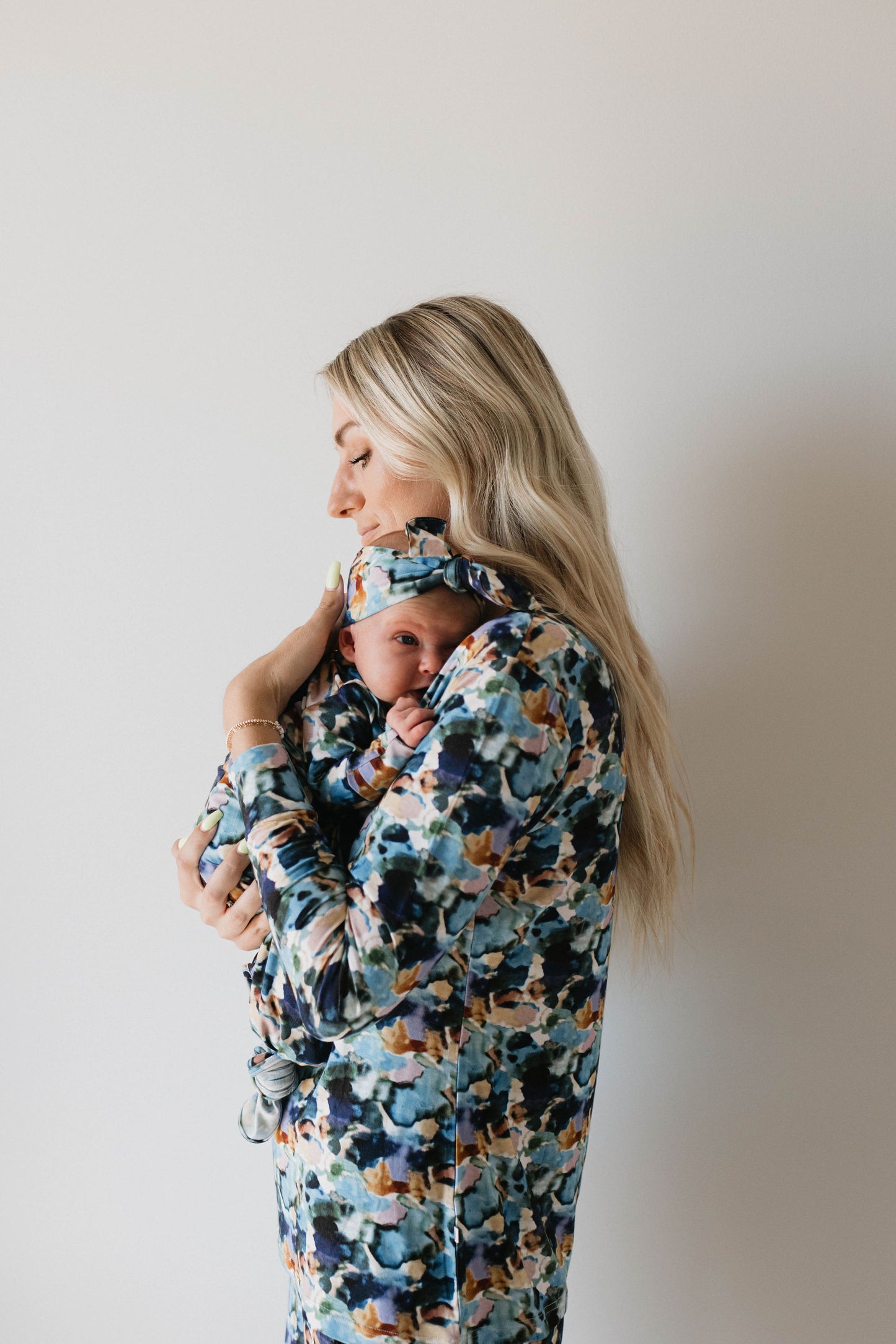 A woman with long blonde hair, wearing a Charli print dress by forever french baby, is cradling a baby wrapped in matching Charli print fabric. She stands against a plain white background, gazing down at the baby with a serene expression. Their outfits remind one of the soft, comforting feel of bamboo pajamas from forever french baby.