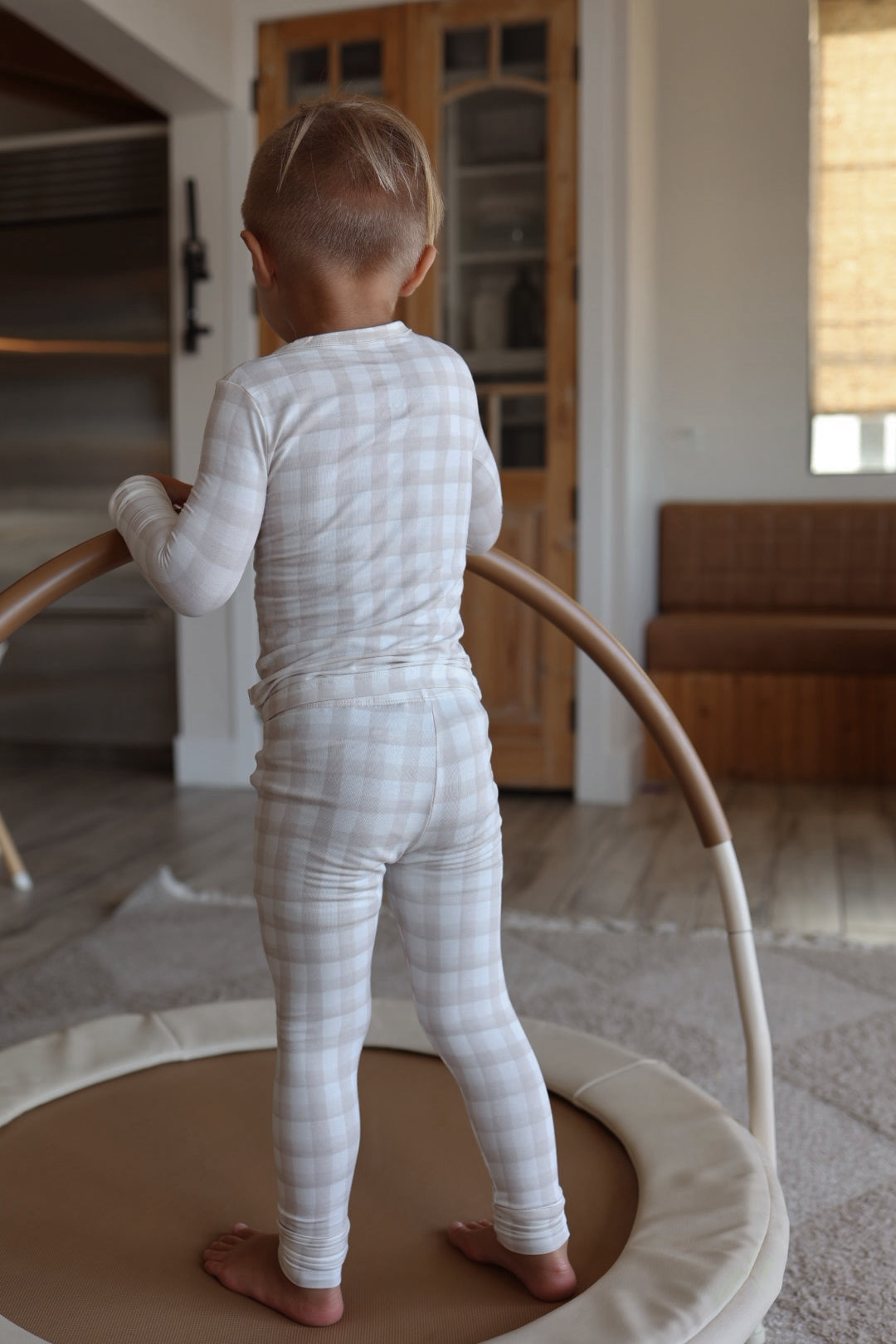 A young child, seen from behind, stands on a padded play mat in forever french baby's Bamboo Two Piece Pajamas | Gingham, holding a circular structure. The room features wooden doors, a couch, and natural light streaming in from the right.