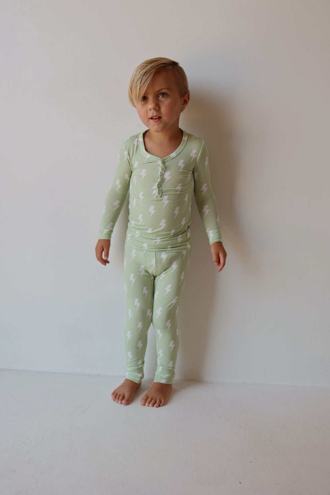 A young child stands barefoot by a plain wall, wearing forever french baby’s Bamboo Two Piece Pajamas | Flash—light green sleepwear with white lightning bolts, made from breathable bamboo for all-night comfort.