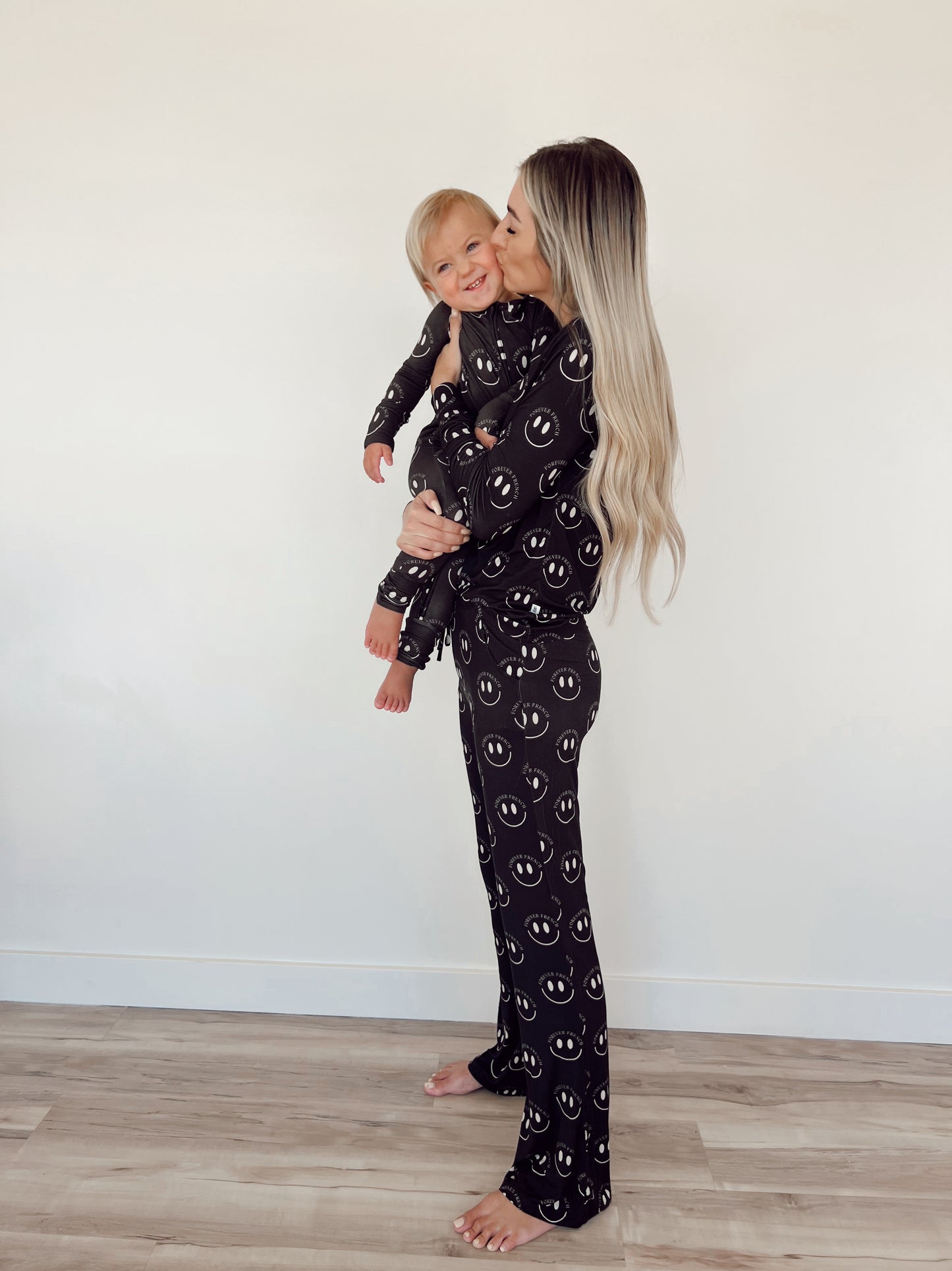 A woman with long, blonde hair stands barefoot on a wooden floor, holding a smiling baby. Both are wearing matching hypo-allergenic pajamas from forever french baby for year-round comfort. The woman is in Women's Bamboo Pajamas in Charcoal & White ff Smile pattern, and she kisses the baby on the cheek against a plain, light-colored wall backdrop.
