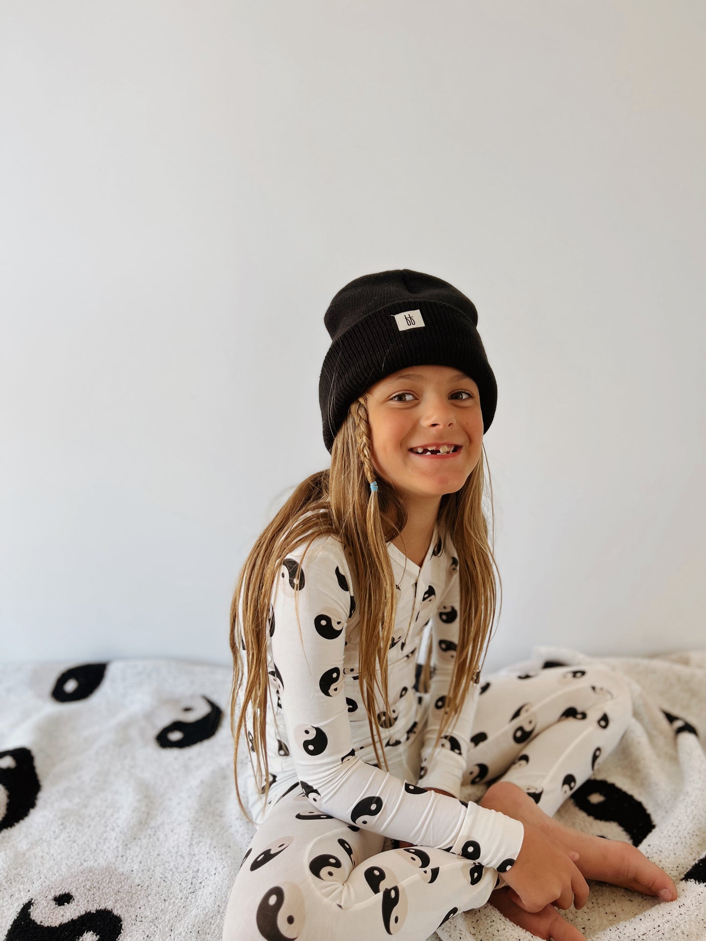 A young child with long hair sits on a bed wearing the Bamboo Two Piece Pajamas | Black & White Yin Yang by forever French baby, adorned with yin-yang symbols. They are also wearing a black beanie and smiling, revealing a gap in their teeth. The bedspread features matching yin-yang patterns, set against a plain white background.