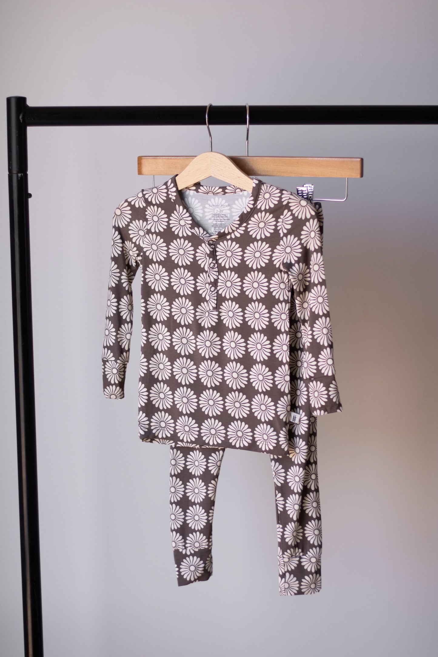 The Bamboo Two Piece Pajamas | Retro Floral by forever french baby features long sleeves and pants with a brown background and white daisy print, hanging on a wooden hanger. Breathable bamboo sleepwear for kids' comfort.