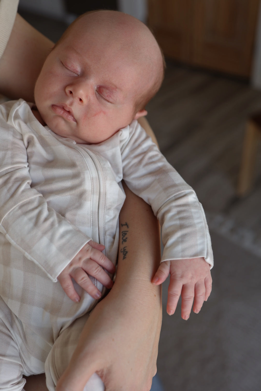 A baby wearing forever french baby's Bamboo Zip Pajamas in Gingham sleeps in someone's arms, their tattooed forearm reading "I love you." A wooden floor and brown door are visible in the background.