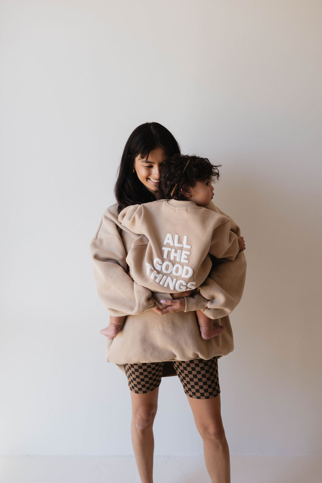 A woman with long dark hair is standing and holding a child. The child is wearing a beige hoodie with the text "ALL THE GOOD THINGS" printed on the back. The woman, dressed in Family Ribbed Biker Shorts in Black & Brown Checkerboard by forever french baby, holds the child's legs wrapped around her waist in a plain room.