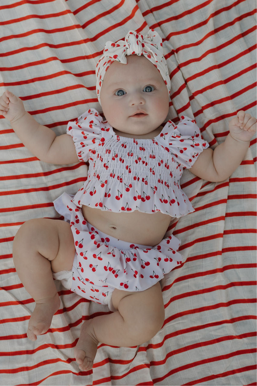 A baby in the forever french baby Toddler Two Piece Swimsuit | Sweetheart Cherries and matching headband lies on a red-and-white striped blanket, gazing up with wide eyes and arms raised.