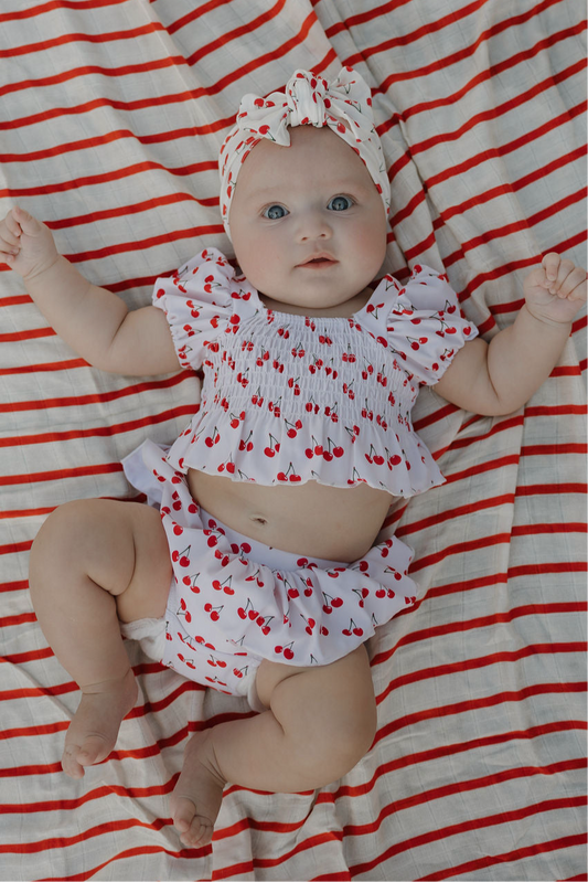 A baby in the forever french baby Toddler Two Piece Swimsuit | Sweetheart Cherries and matching headband lies on a red-and-white striped blanket, gazing up with wide eyes and arms raised.