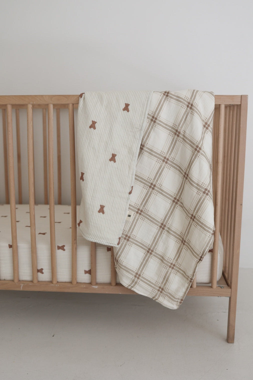 A wooden baby crib with a white, bear-print mattress is topped by the Muslin Quilt | Teddy & Ellis Plaid from forever french baby, draped over the rail. The background wall is plain white.