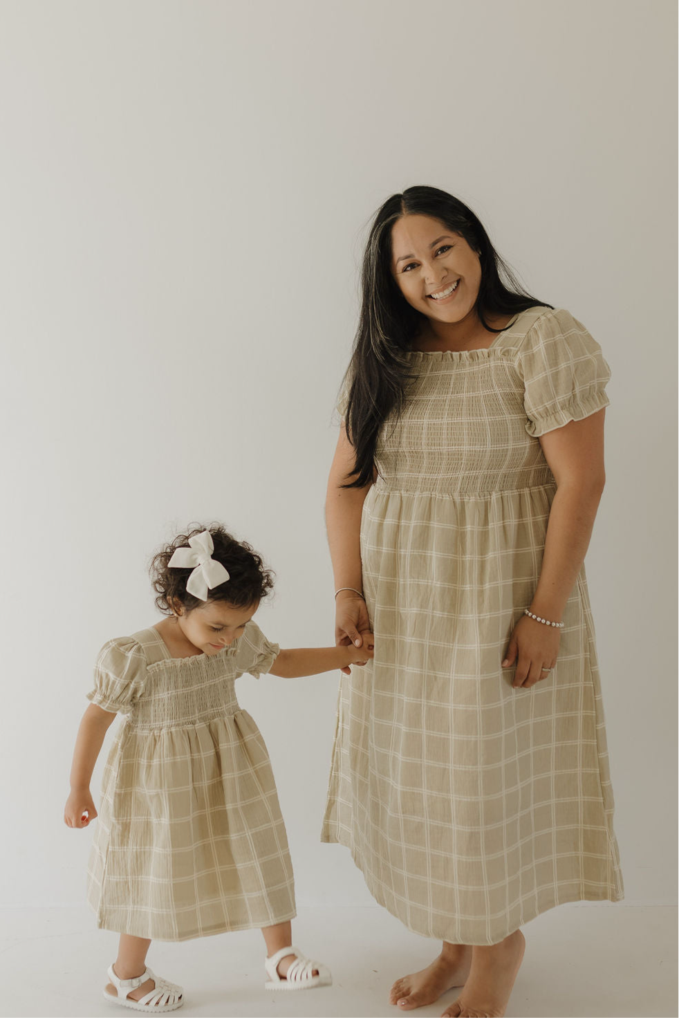 A woman and young girl, both barefoot against a light background, hold hands and smile in matching forever french baby Women's Short Sleeve Muslin Dresses (Linen Grid); the girl's curly hair features a white bow.