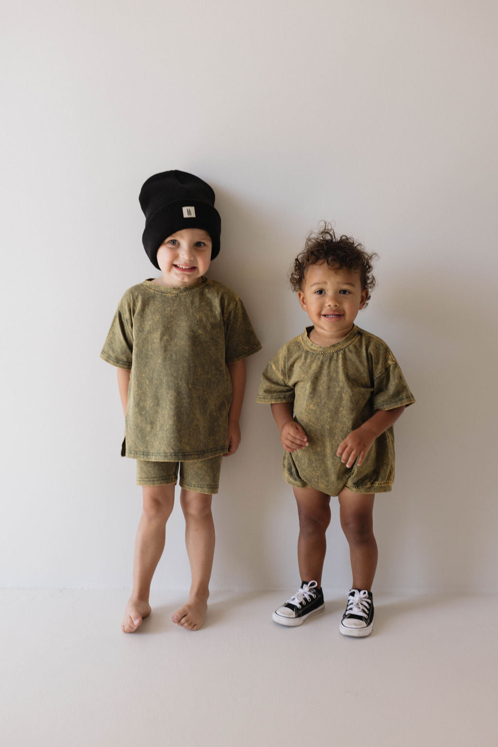 Two young children stand against a plain white background. The older child, barefoot and wearing an ff Knit Beanie in black, is dressed in a greenish-brown T-shirt and shorts. The younger child, wearing a ribbed design onesie and black shoes from Forever French Baby, smiles brightly alongside their sibling.