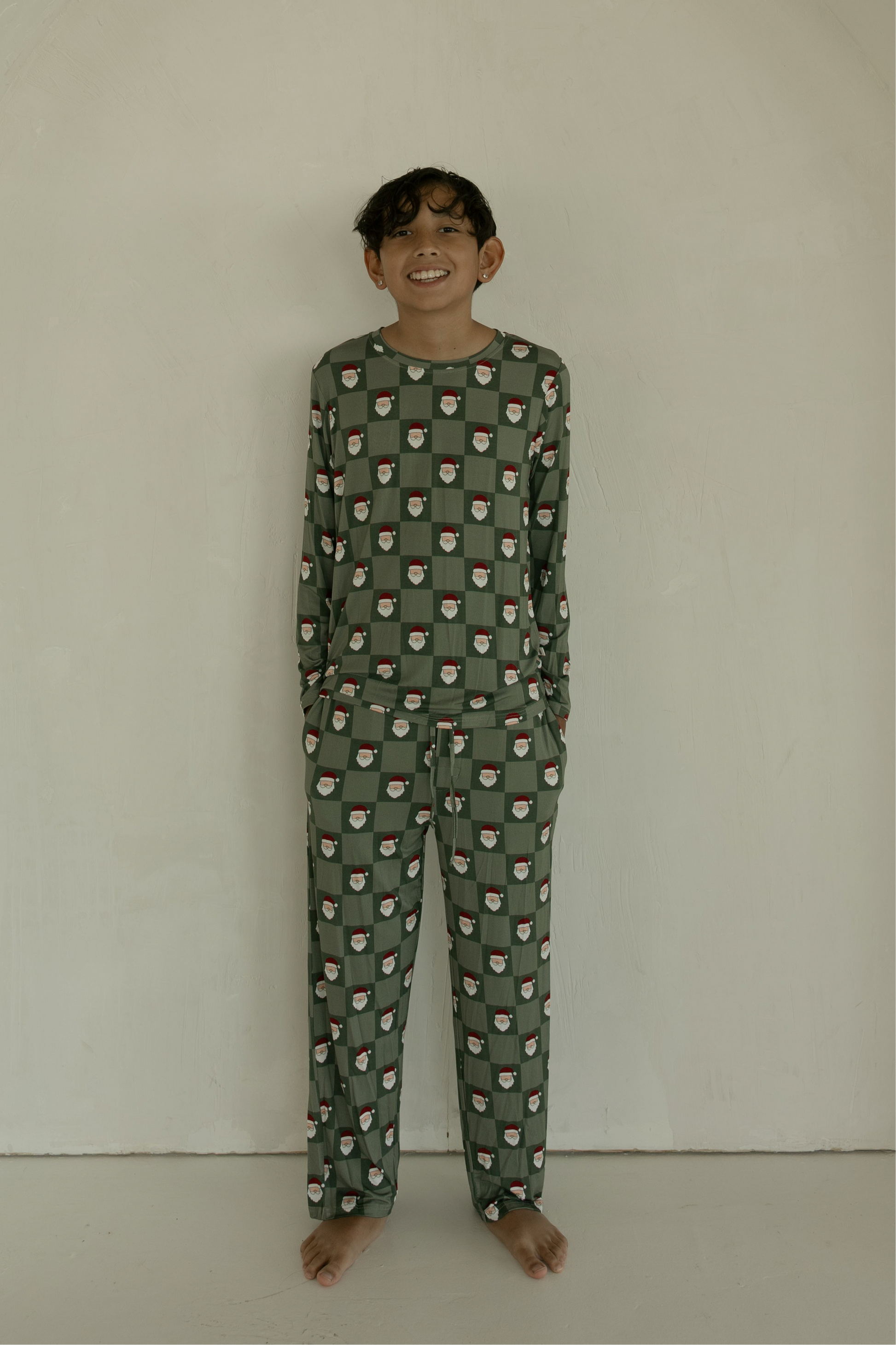 A smiling, dark-haired child stands barefoot against a light wall, wearing forever french baby’s Pre-Teen Straight Leg Bamboo Pajamas in the Santa Checkerboard print, with hands in their pockets.