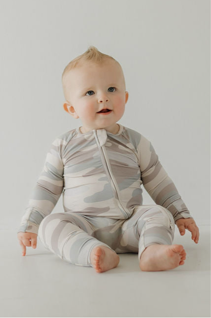 A light-haired baby sits on the floor in forever french baby Bamboo Zip Pajamas | Camo, a breathable, camouflage-patterned zip-up onesie, looking curiously upward against a plain white background.