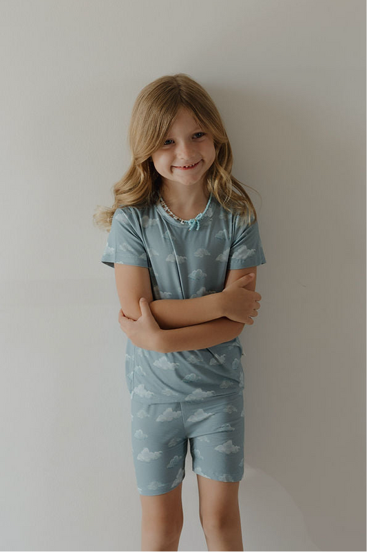 A young girl with long blonde hair smiles against a plain wall, wearing the Kendy 3 Bamboo Short Set | Duke—blue hypo-allergenic pajamas with a white cloud pattern, offering cozy, breathable comfort perfect for sensitive skin.