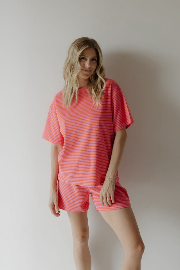 A woman with long blonde hair wears the forever french baby Women's Terry Short Set in Strawberry, featuring a loose fit and short sleeves, as she stands against a plain light wall, looking at the camera and smiling softly.