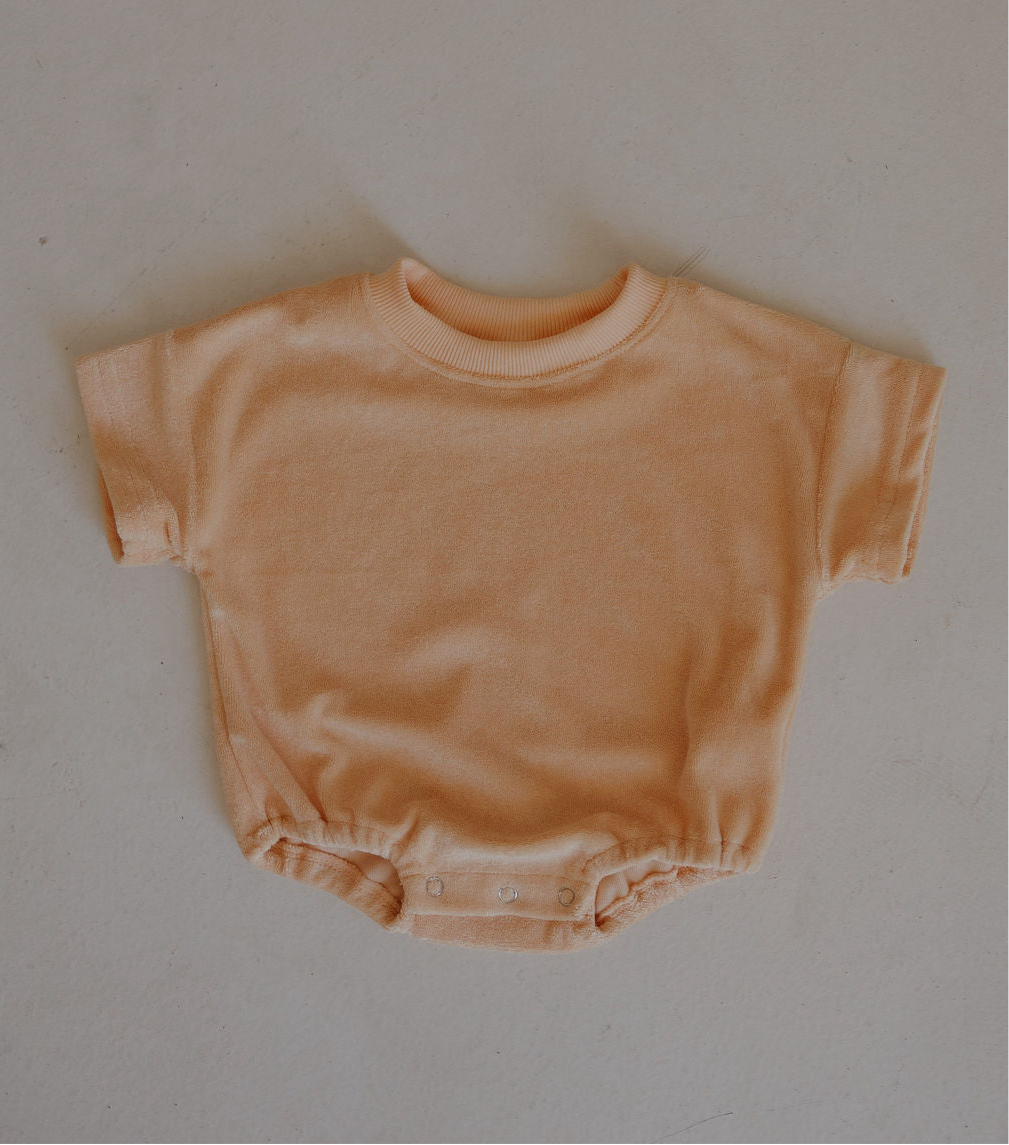 The forever french baby Terry T-Shirt Romper | Sherbert is a peach-colored, short-sleeve, terry romper with a round neck and snap buttons at the crotch for easy diaper changes, shown laid flat on a light surface.