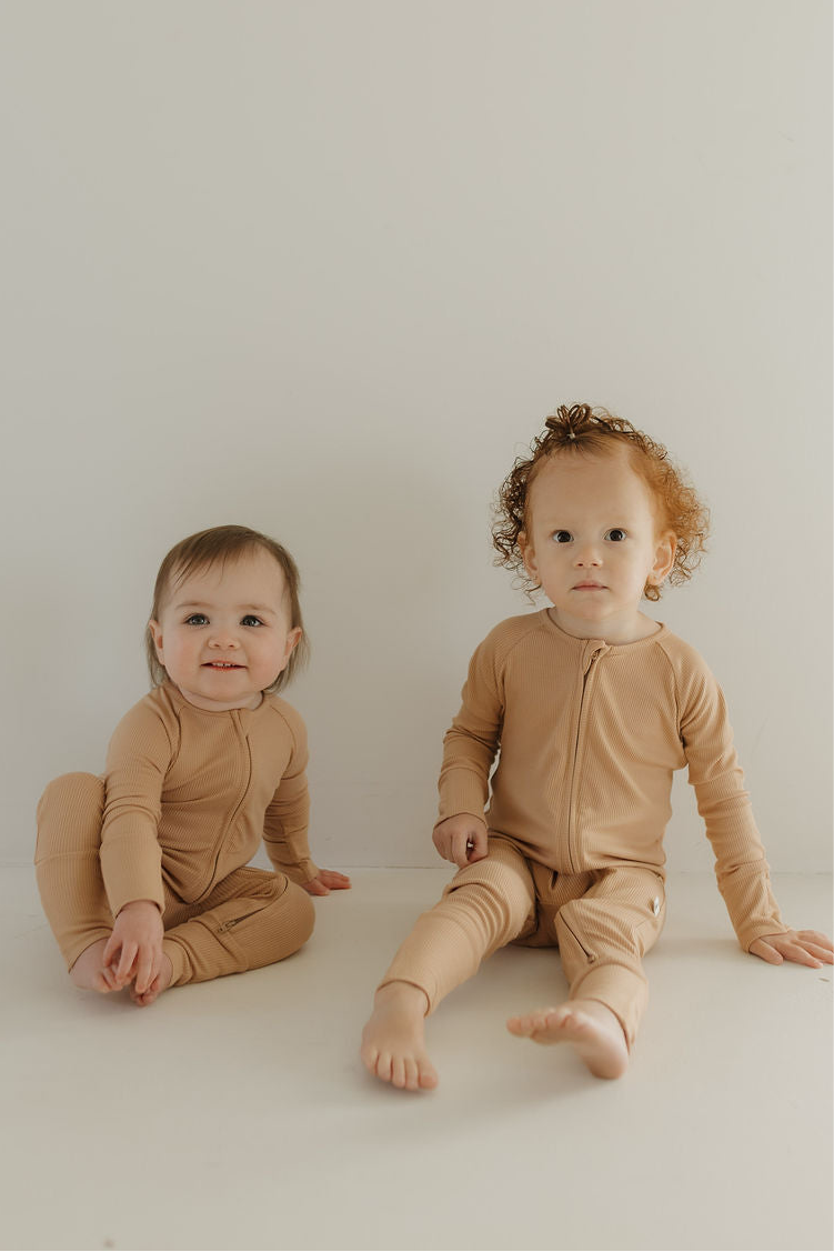 Two toddlers sit by a white wall in matching forever french baby Bamboo Zip Pajamas | Tawny. One smiles with crossed legs, the other sits with legs extended—soft bamboo comfort and style, perfect for all seasons.