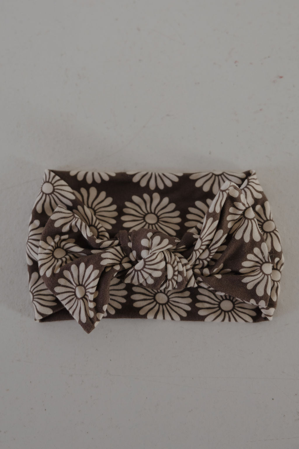 The Bamboo Head Wrap | Retro Floral by forever french baby, a small rectangular item wrapped in brown fabric with white daisies and tied in a neat bow, is displayed on a plain light surface.