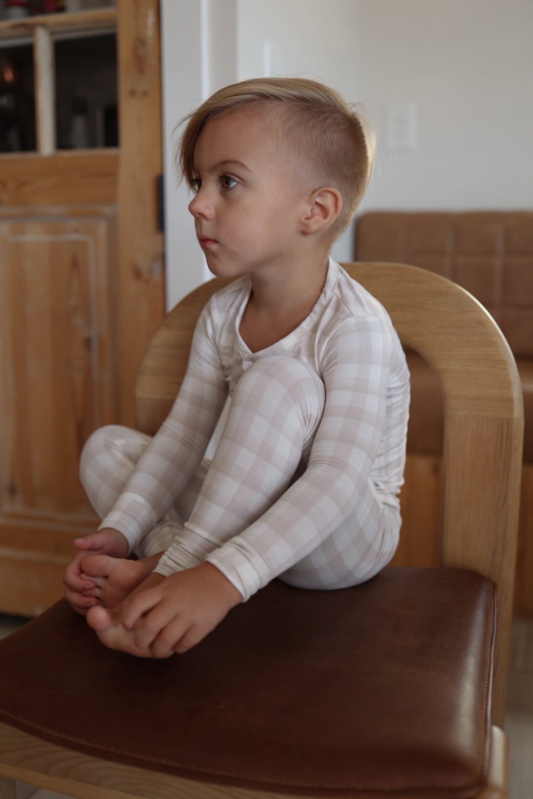 A young child with short hair sits cross-legged on a wooden chair, wearing forever french baby’s Bamboo Two Piece Pajamas | Gingham, looking thoughtfully to the side in a cozy room.