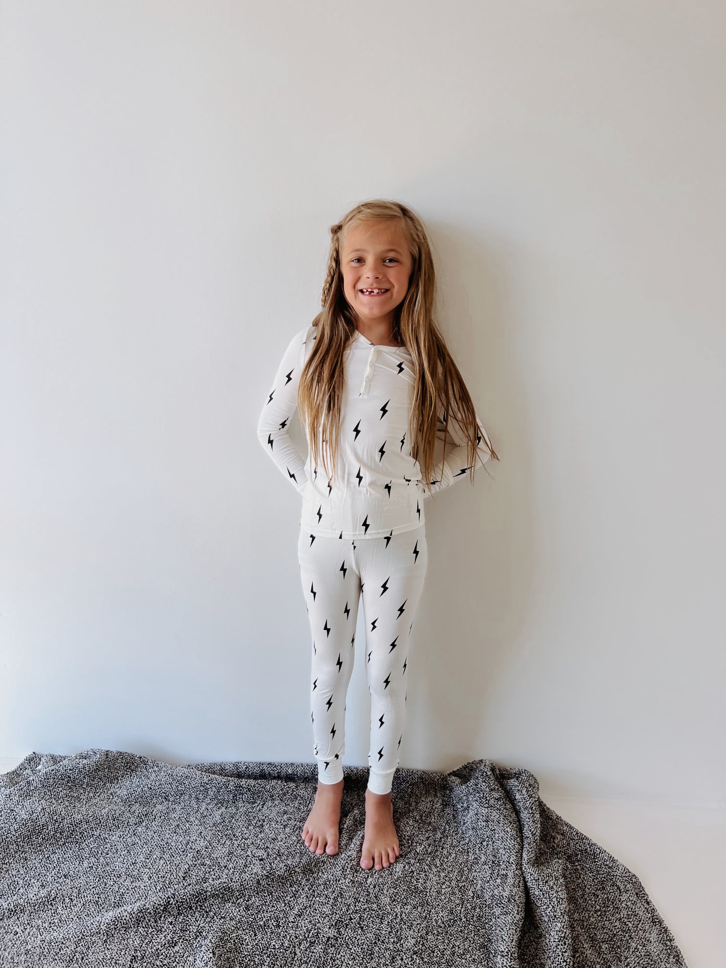 A young girl with long hair stands barefoot on a gray rug against a plain white wall. She is wearing forever french baby's Bamboo Two Piece Pajamas in Black & White Lightning Bolt, made from hypo-allergenic bamboo clothing. She smiles, with her hands resting on her sides.