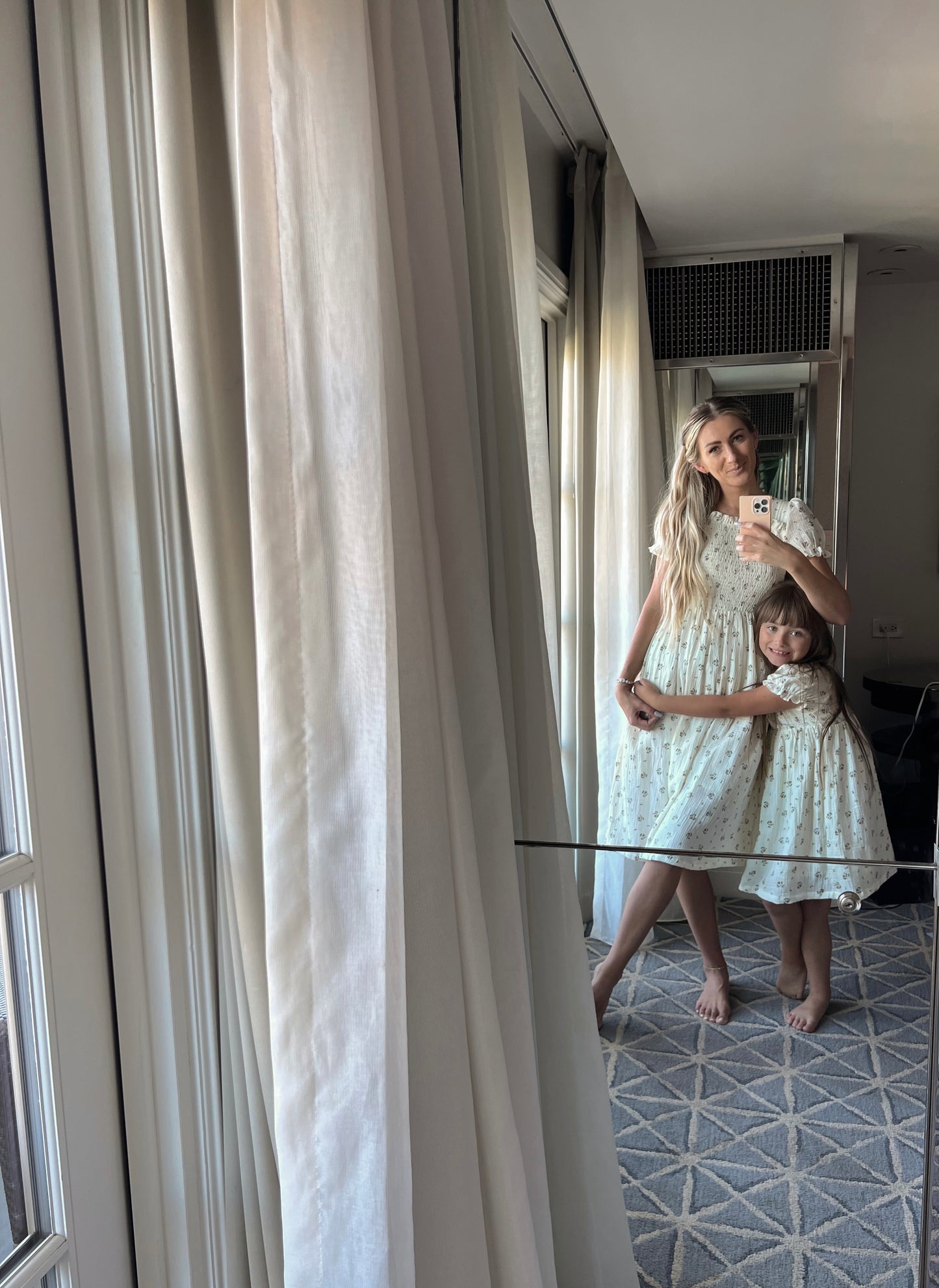 A woman and a young girl, both dressed in forever french baby's Garden Days Child & Women's Muslin Dresses in light-colored floral prints, stand barefoot in front of a mirror. The woman holds her phone to take a selfie while the girl hugs her around the waist. They are standing on a patterned carpet with curtains beside them.