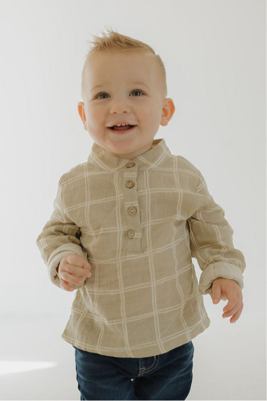A smiling toddler with short blonde hair wears the forever french baby Muslin Button Up | Linen Grid shirt and dark jeans, standing against a light background.