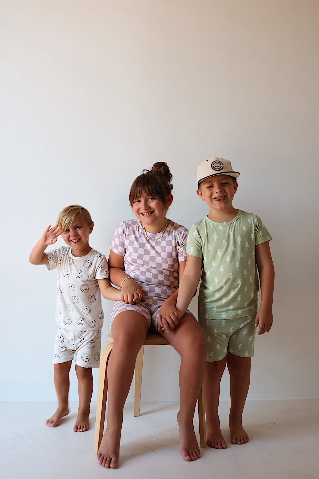 Three young children smile against a plain backdrop. One sits on a stool while the others stand nearby, all dressed in forever french baby’s Bamboo Short Set | Green Smiles—breathable sleepwear pajamas combining comfort and style.