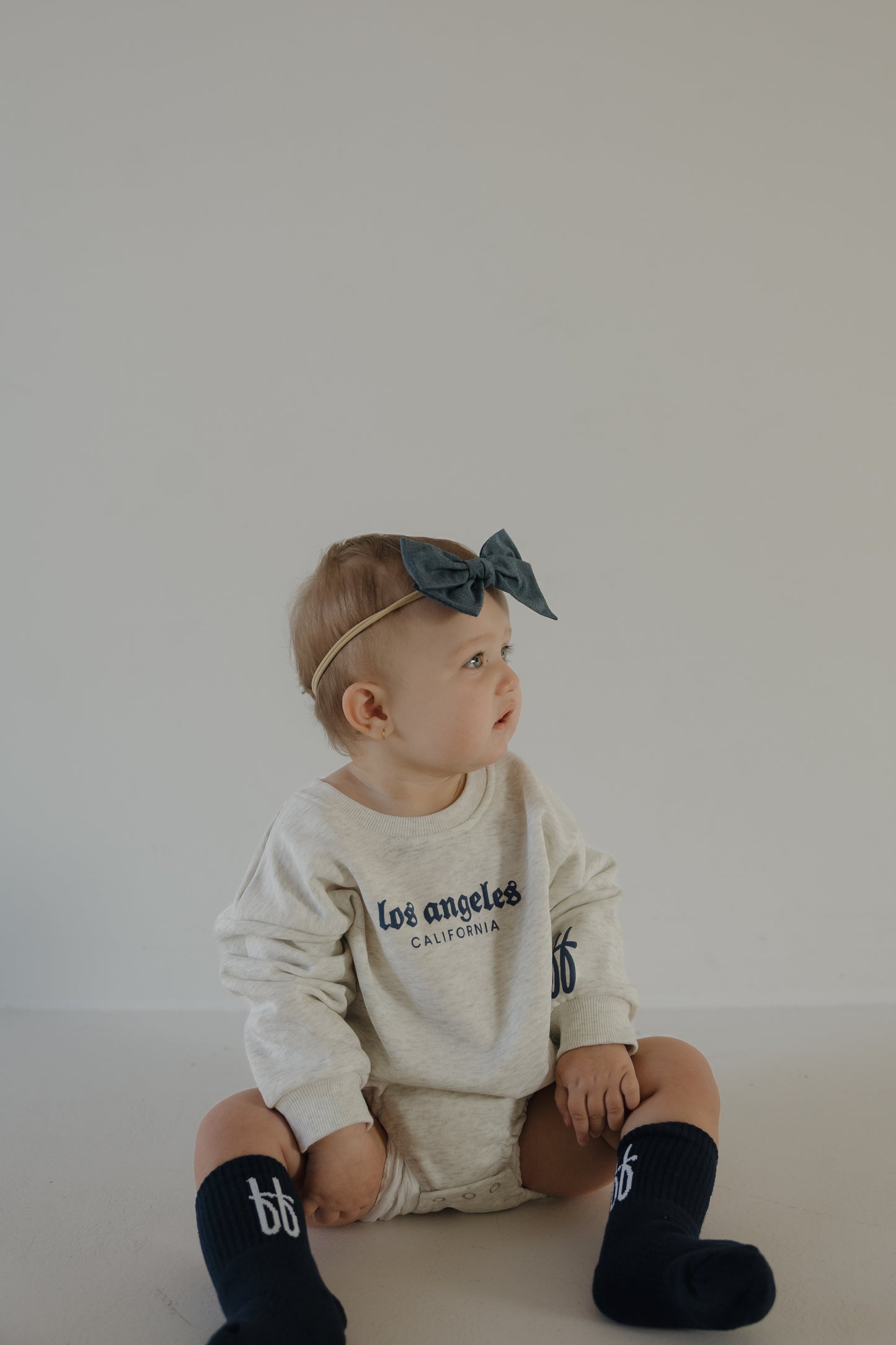 A baby wearing a blue headband, navy blue socks, and the forever french baby Sweatshirt Romper | ff Los Angeles sits on a light floor against a plain, light-colored wall, looking to the side.