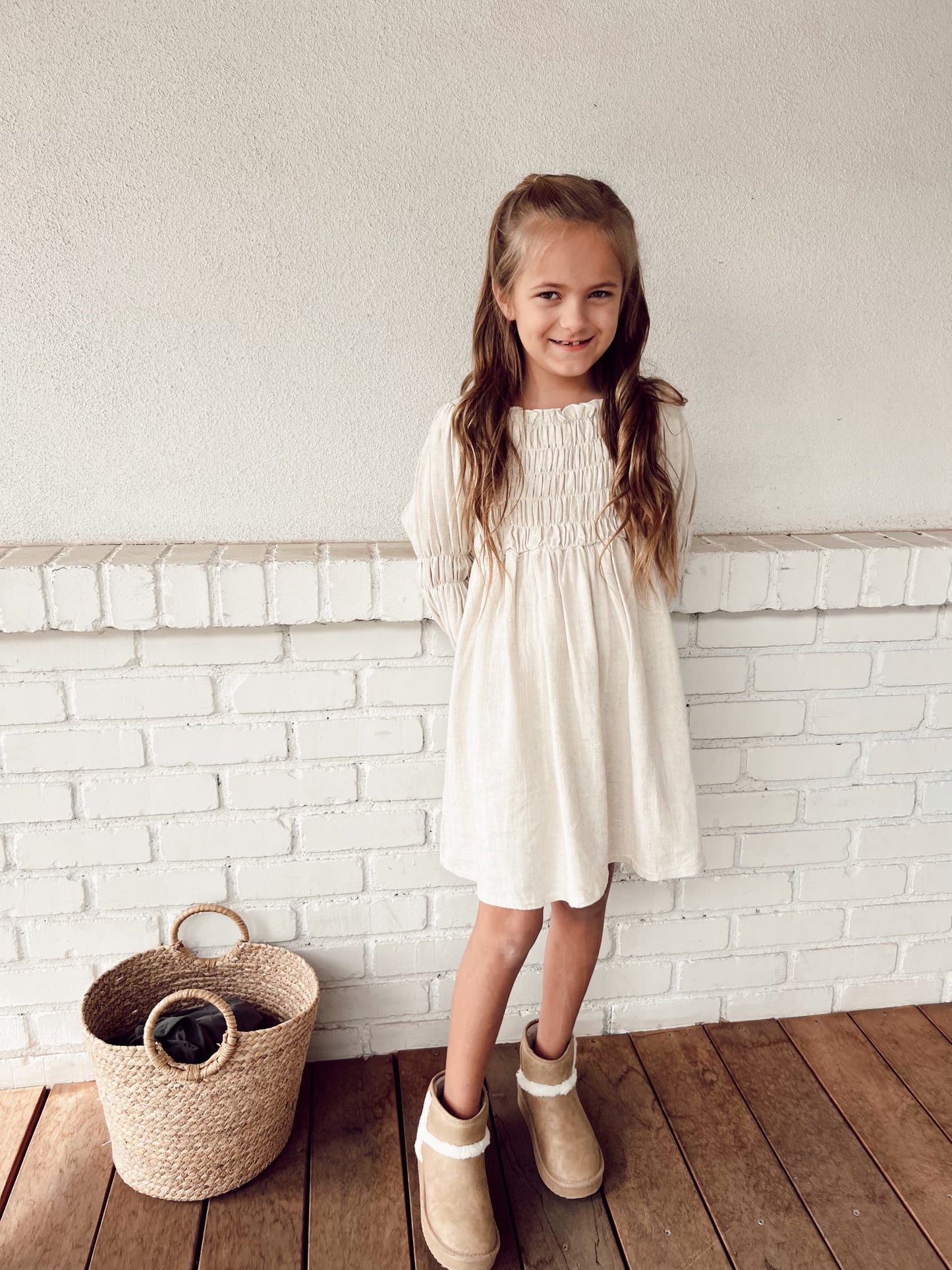 A young girl with long hair stands on a wooden floor in front of a white brick wall, wearing the Women's Linen Dress | Flax from forever french baby and brown boots. Her hands are behind her back, and there is a woven basket to her left.