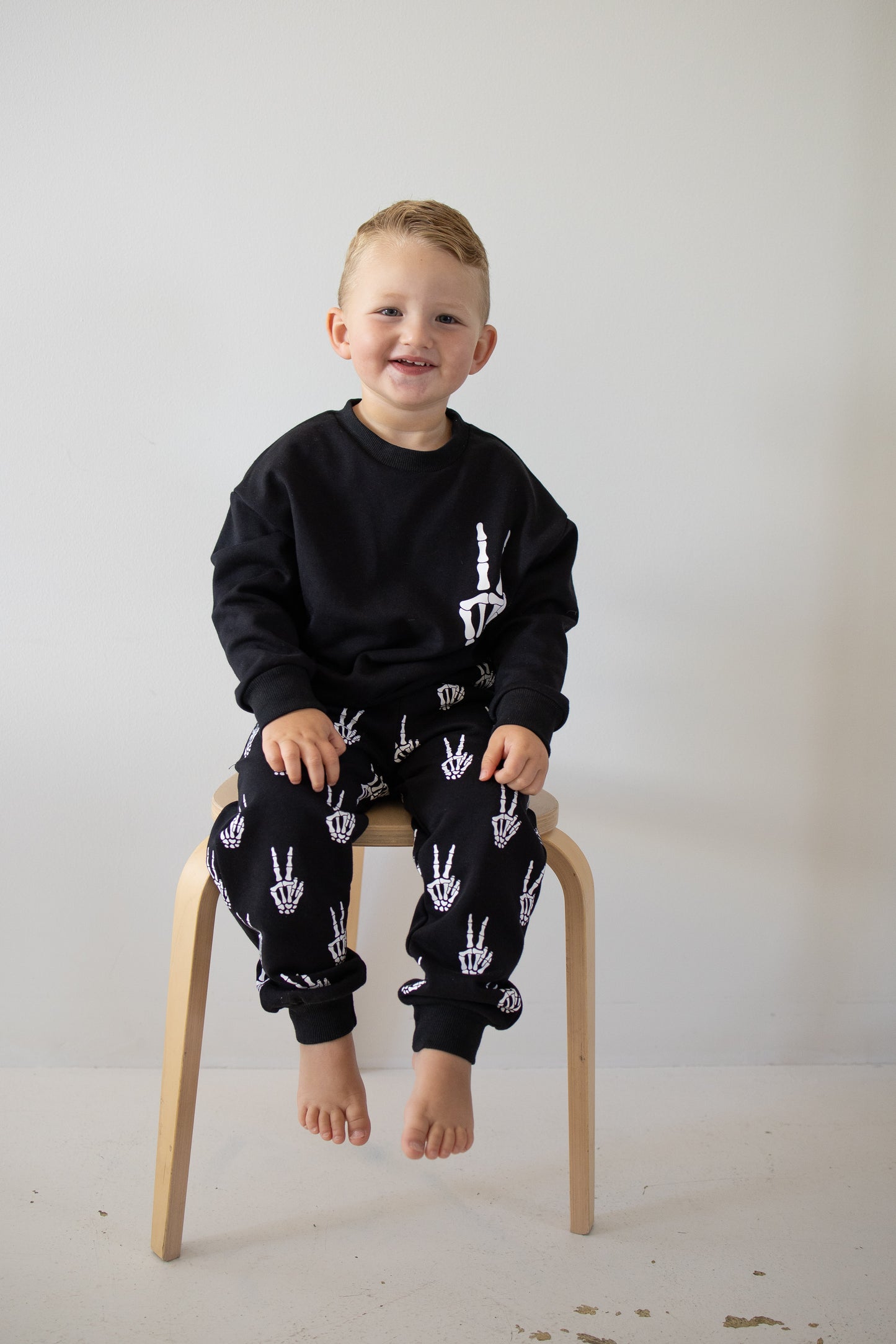 A young child with light hair sits barefoot on a wooden stool, wearing the forever french baby Child Sweat Set | Midnight Skelly, featuring playful white hand and candle designs—perfect for everyday adventures—against a plain light-colored wall.