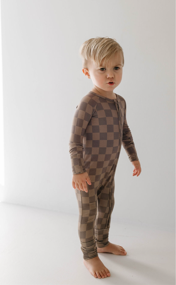 A young, barefoot child with blonde hair stands on a light floor, wearing forever french baby's Bamboo Zip Pajamas in Faded Brown Checkerboard and looking slightly to the side.