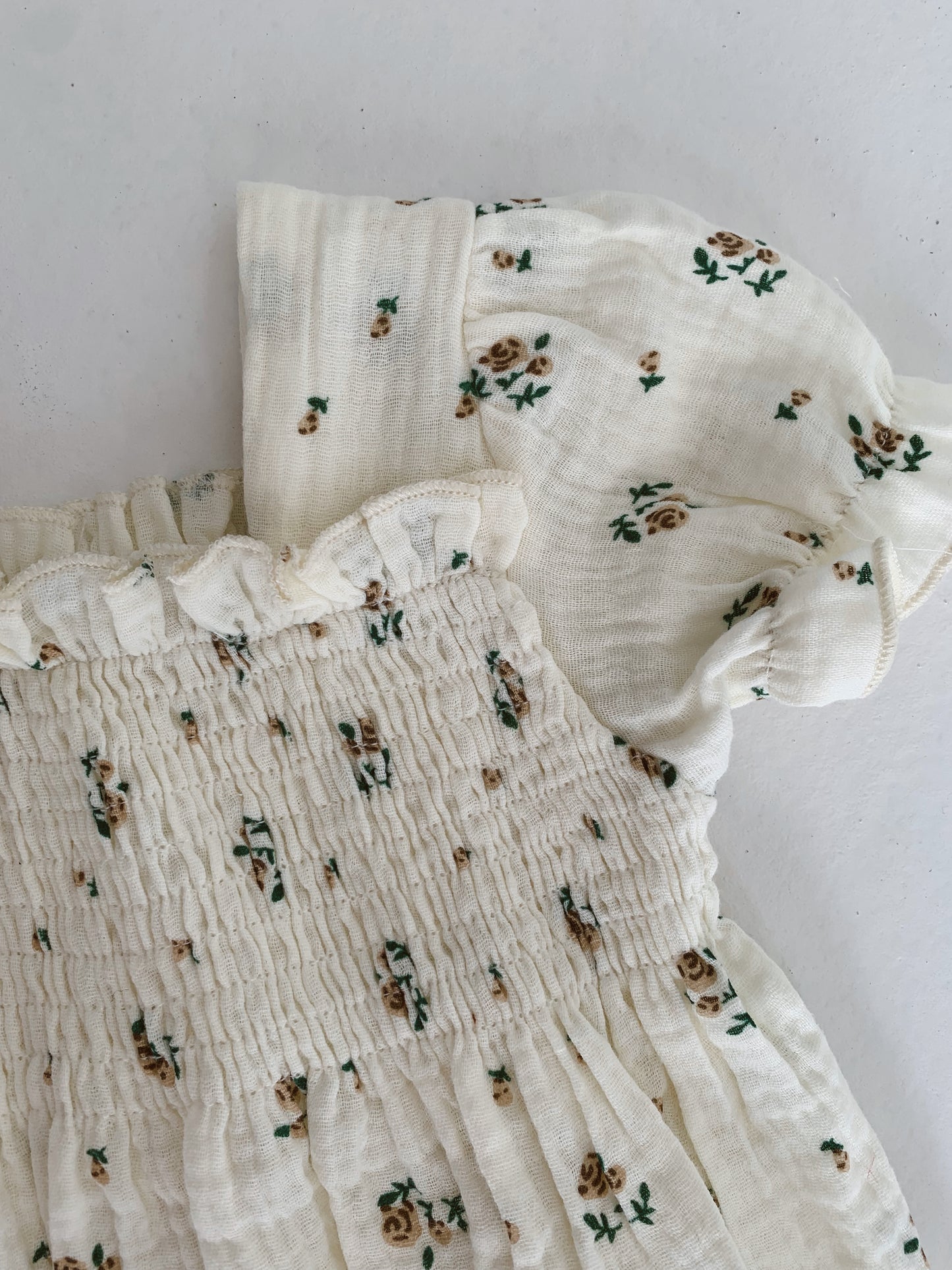 A close-up of the "Garden Days" muslin dress by forever french baby, showcasing its ivory fabric adorned with small brown floral prints. The dress features smocked detailing at the bodice and short, puffed sleeves. Crafted from light, textured material, it's perfect for warm weather and ideal for matching mom-and-daughter outfits.