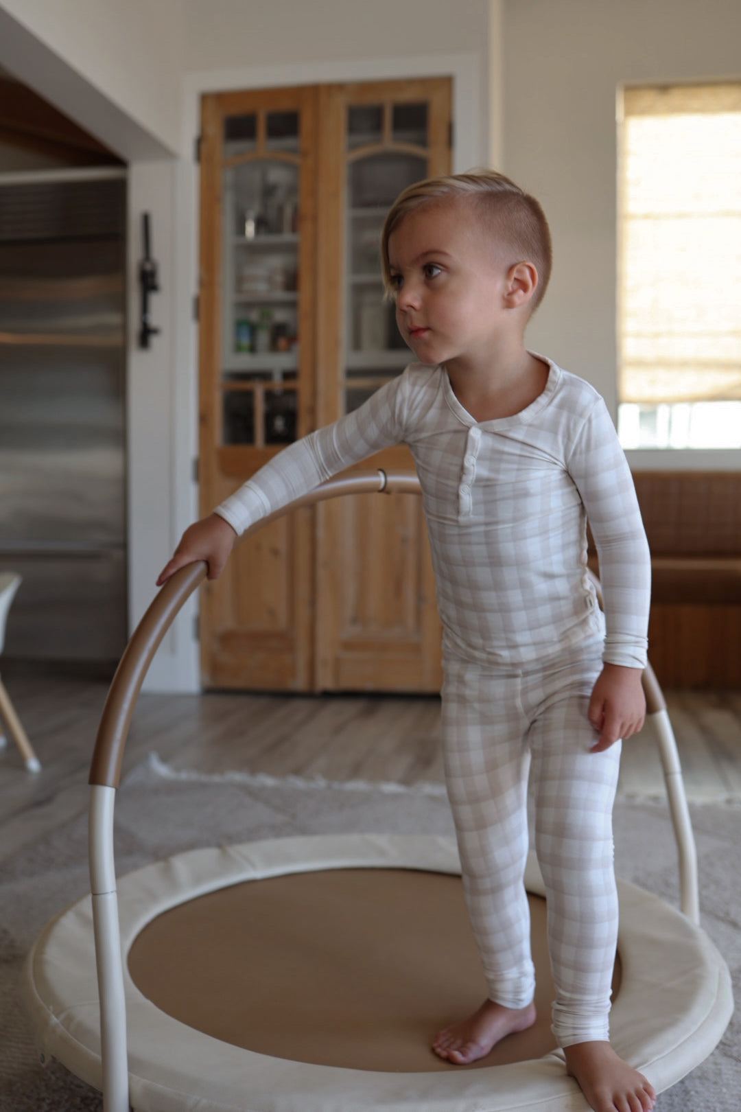 A young child in forever french baby's Bamboo Two Piece Pajamas | Gingham stands barefoot on a round indoor play trampoline, holding the curved handle in a cozy, modern living room with wooden doors and neutral tones.