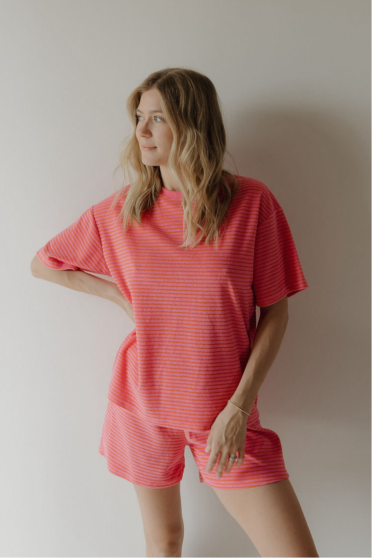 A woman with wavy blonde hair stands against a plain wall, wearing the forever french baby Women's Terry Short Set in Strawberry, perfectly showcasing the stylish rompers and sets trend with one hand on her hip.